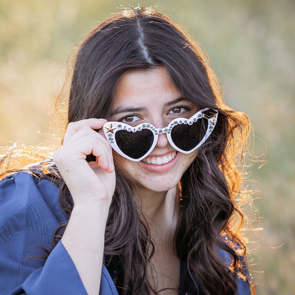 Young woman smiling and holding heart-shaped sunglasses with floral frames outdoors in warm golden light.