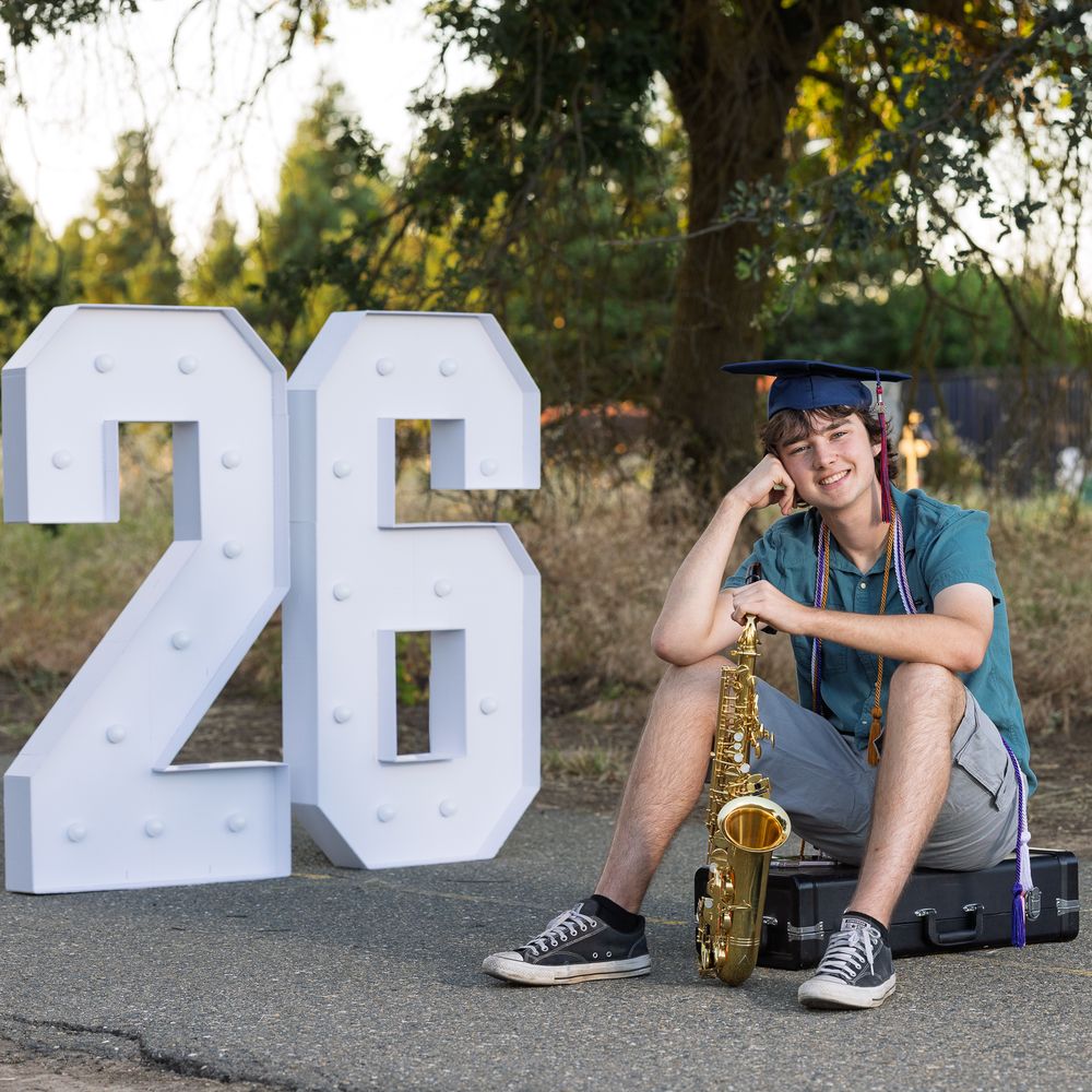 Teen graduate in cap poses with saxophone beside illuminated number 26 sign outdoors.