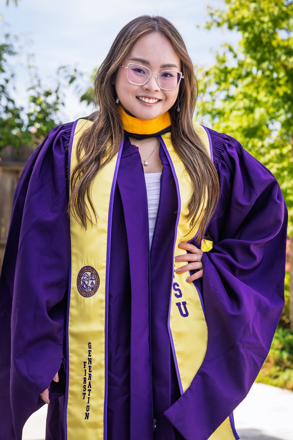Smiling graduate wearing purple and gold graduation gown and stole, posing outdoors with trees in background.