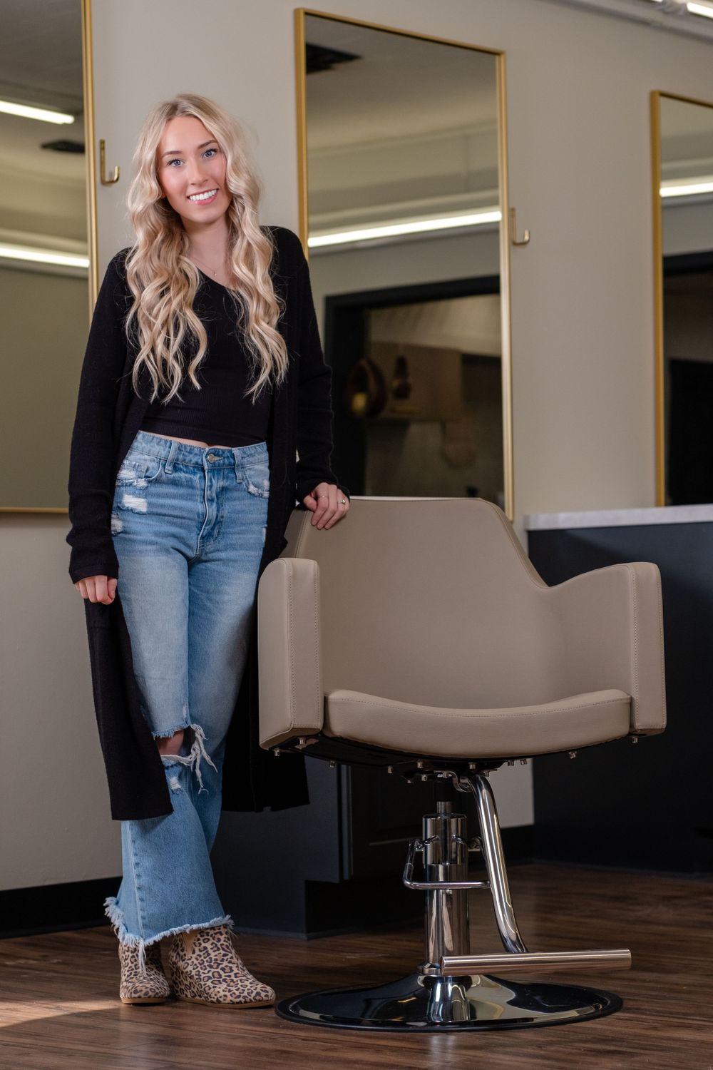 A woman in ripped jeans and a black sweater stands next to a salon styling chair on a hardwood floor.