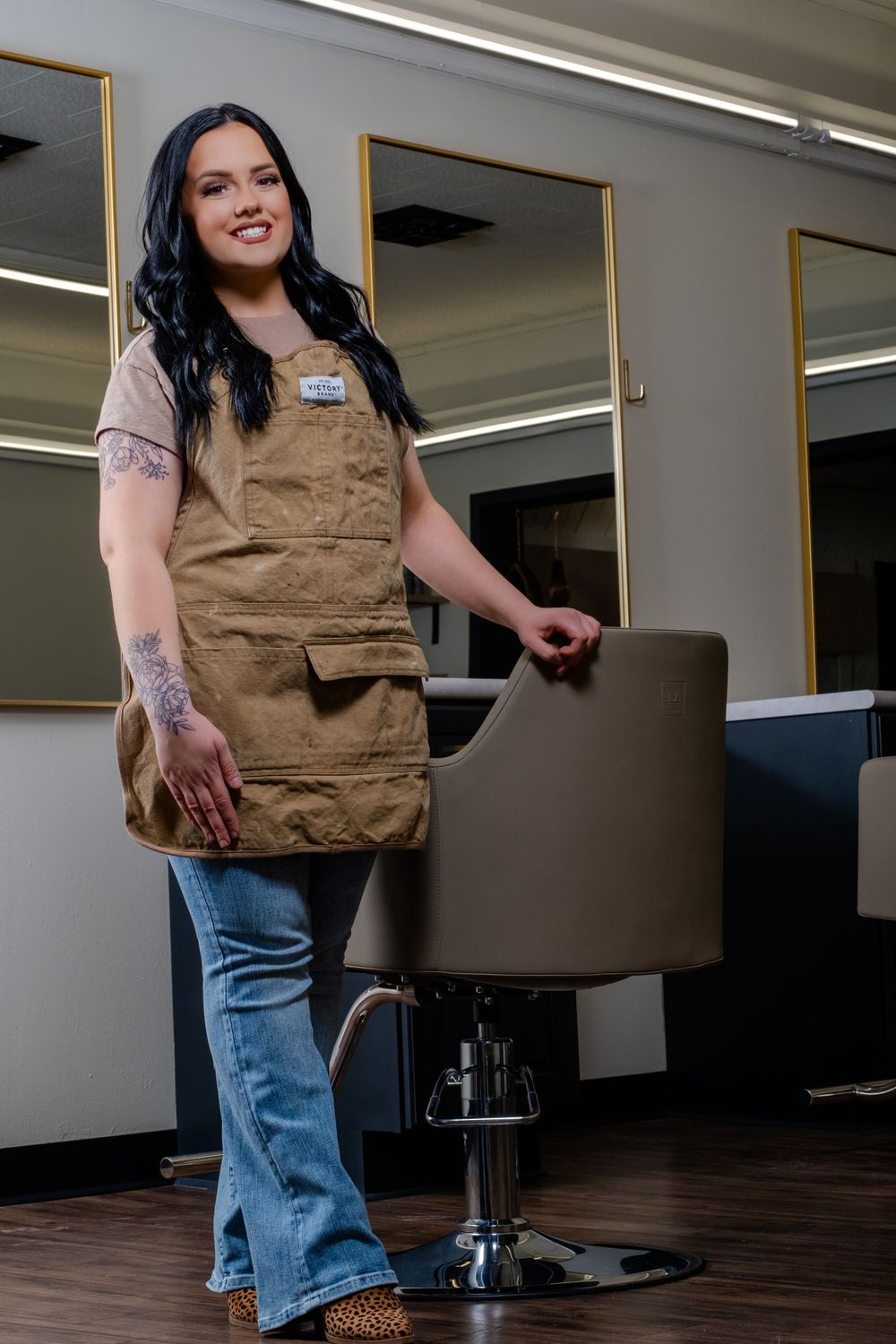 A hairstylist wearing a brown apron and jeans stands confidently next to a salon chair in a modern salon setting.