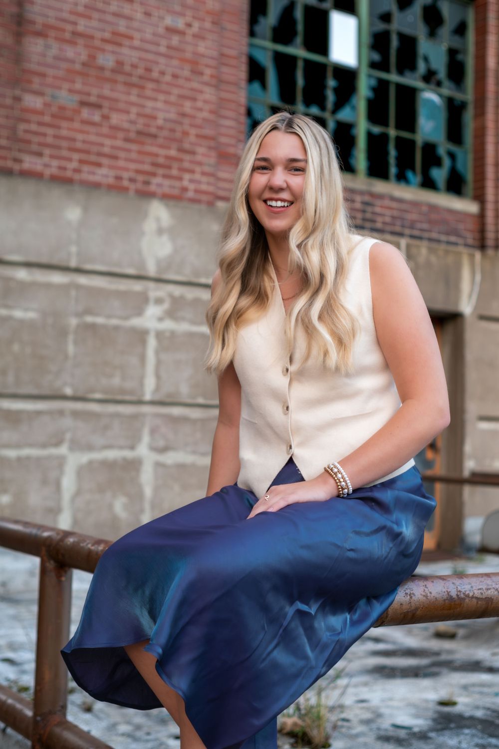 Smiling person in cream sleeveless top and navy skirt sits casually on bench against brick wall.
