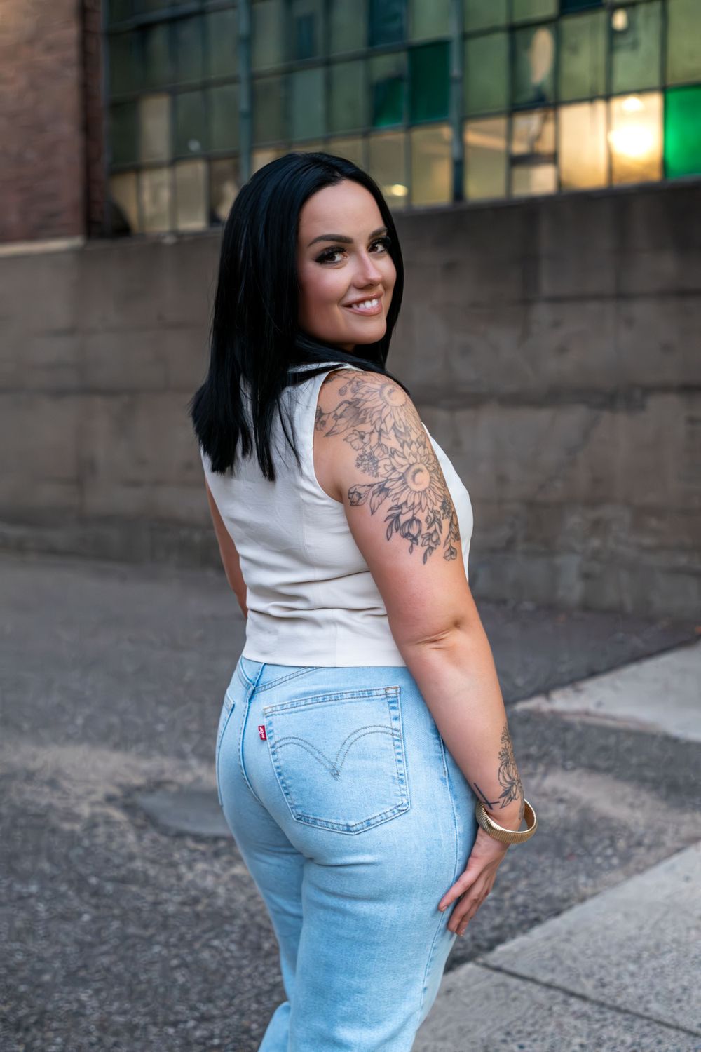 A woman in a white tank top and light jeans poses on a city street, showing off her arm tattoo at sunset.