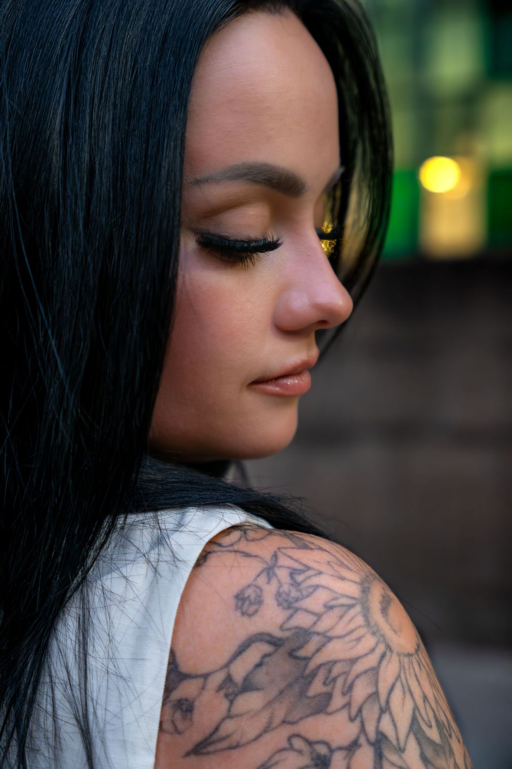 A close portrait of a person with closed eyes shows off their detailed shoulder tattoo in soft lighting.