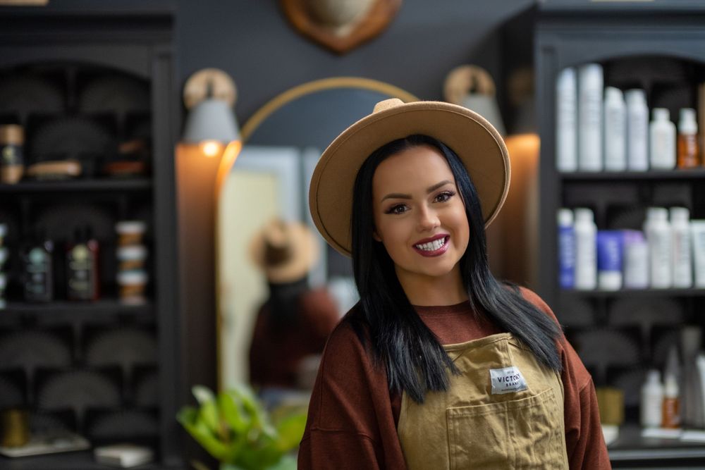 Cheerful stylist wearing a wide-brimmed hat and brown apron stands inside a salon with shelves of products behind them.