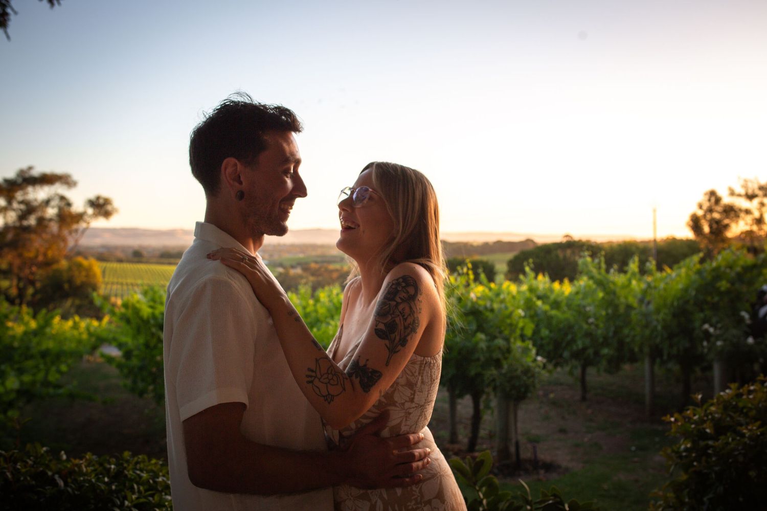 Romantic couple embraces in a vineyard at sunset with golden light filtering through the vines.
