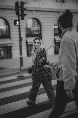 A couple crossing a city street, one wearing a leopard print coat while looking back with joy.