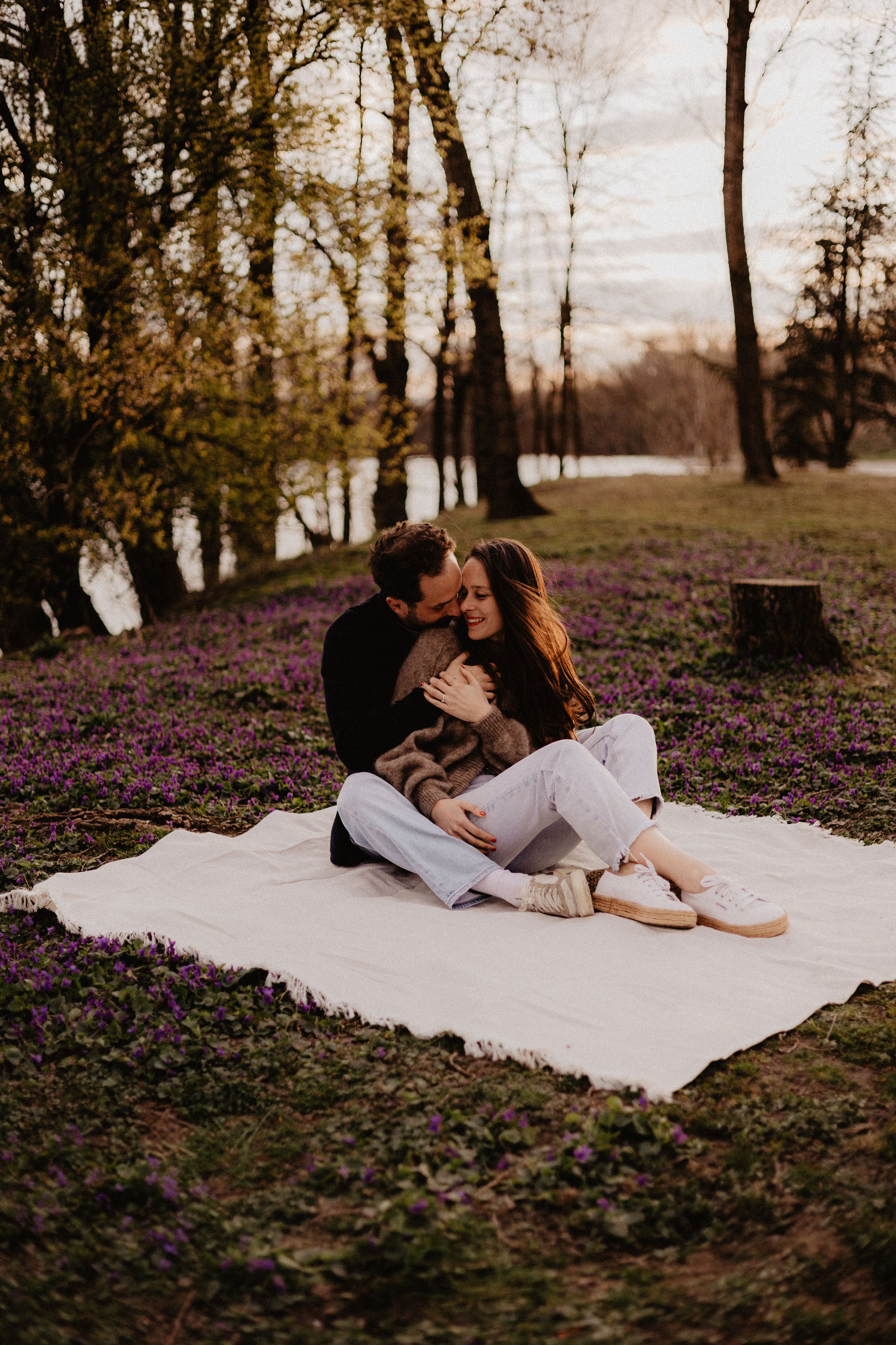 A romantic couple lying on a white blanket surrounded by purple wildflowers in a dreamy outdoor setting.