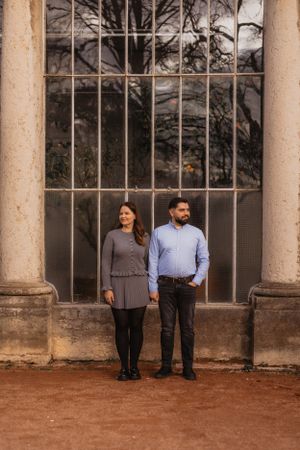 Couple in formal attire standing together against a rustic greenhouse backdrop during a photo session.