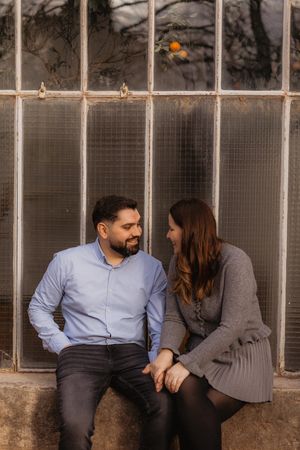 Couple sitting close together on stone ledge in front of greenhouse windows during outdoor photoshoot.
