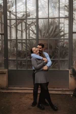 Playful couple embracing and laughing together in front of greenhouse windows during photoshoot.