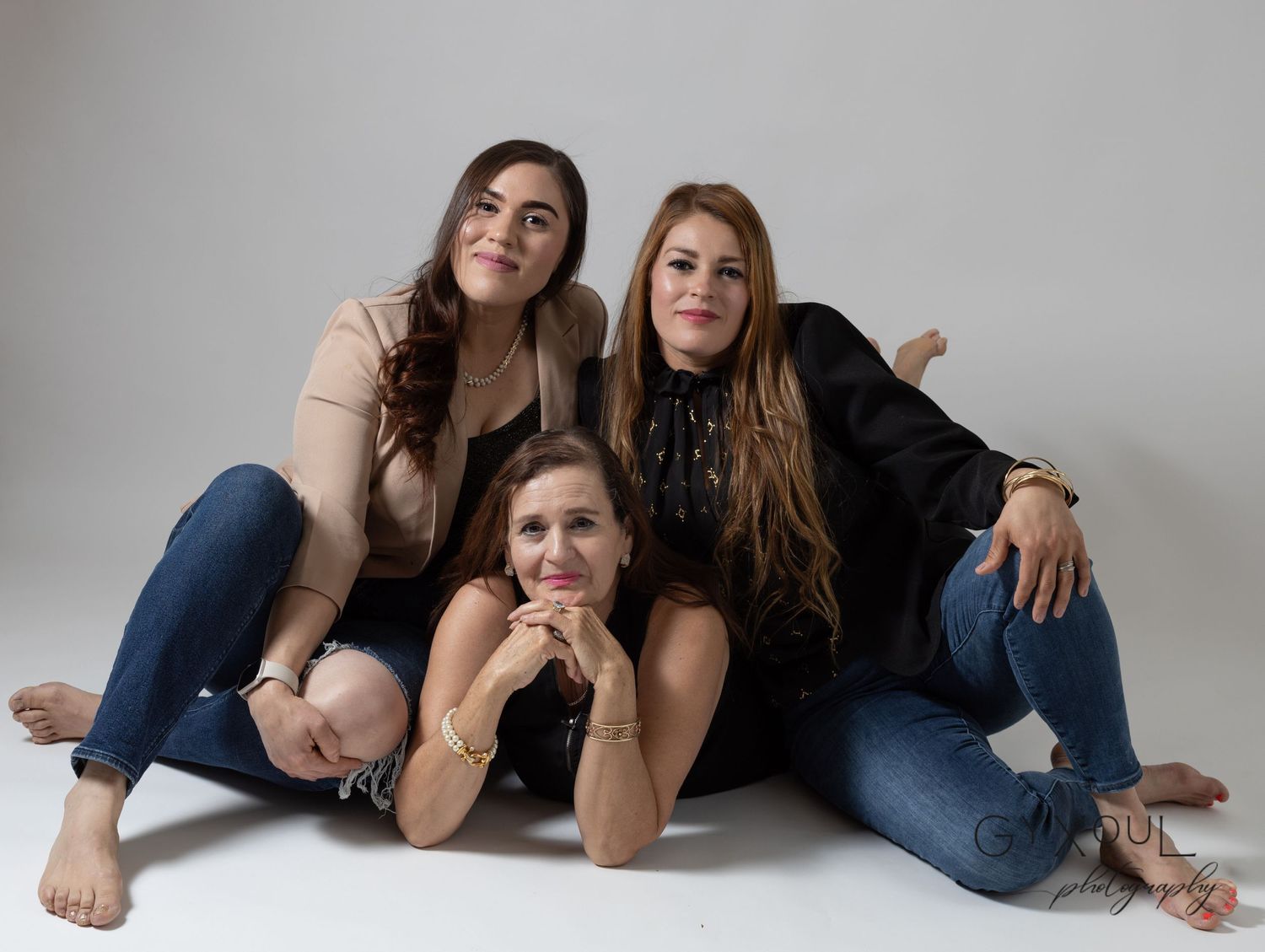 Three women in casual attire pose together on the floor in a relaxed studio portrait.