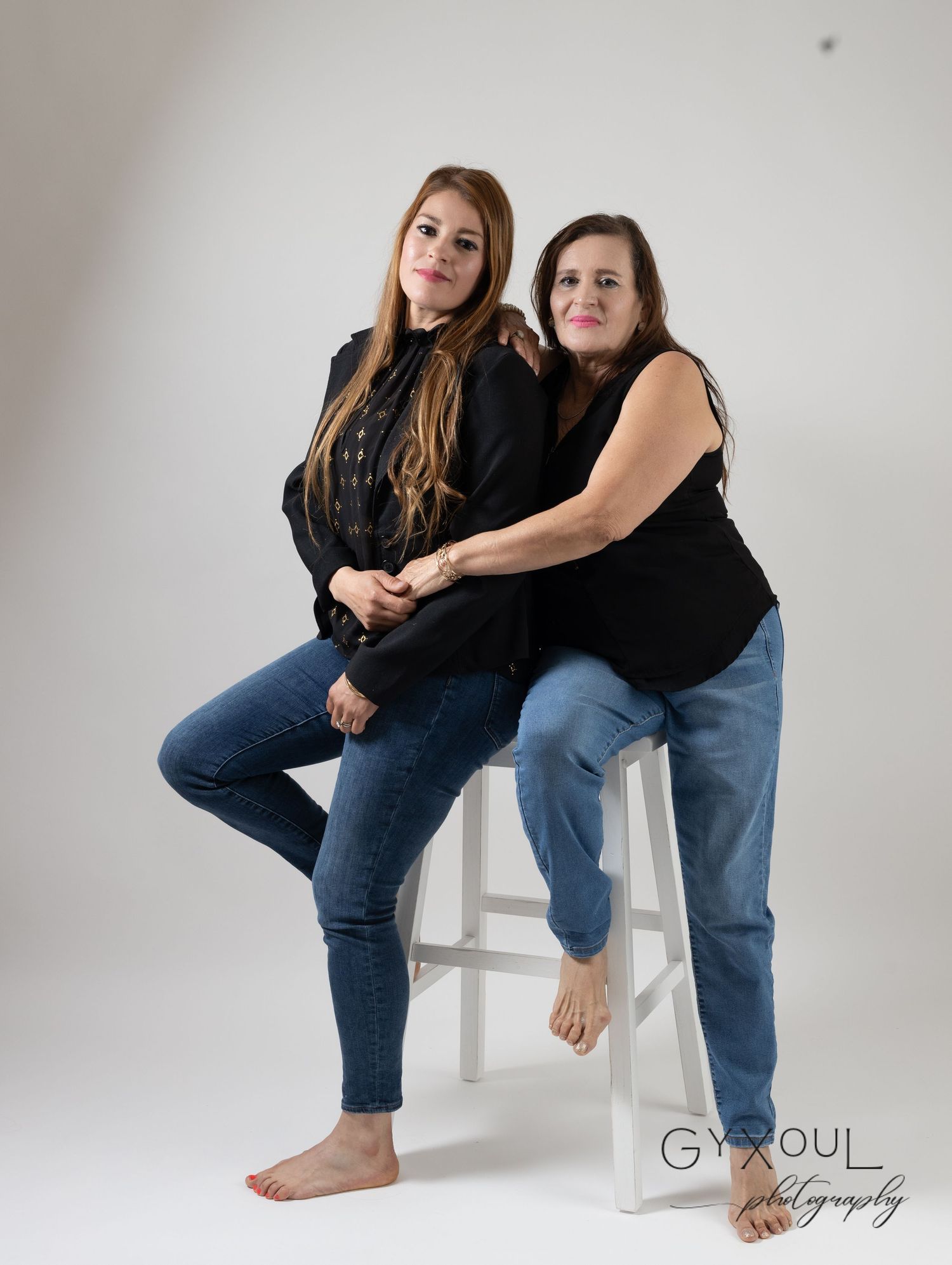 Two women in black tops and jeans pose casually on a white stool against a studio backdrop.