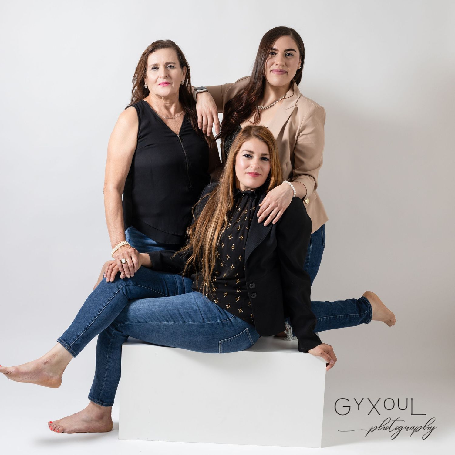 Three women in casual outfits pose together on a white cube in a studio setting.
