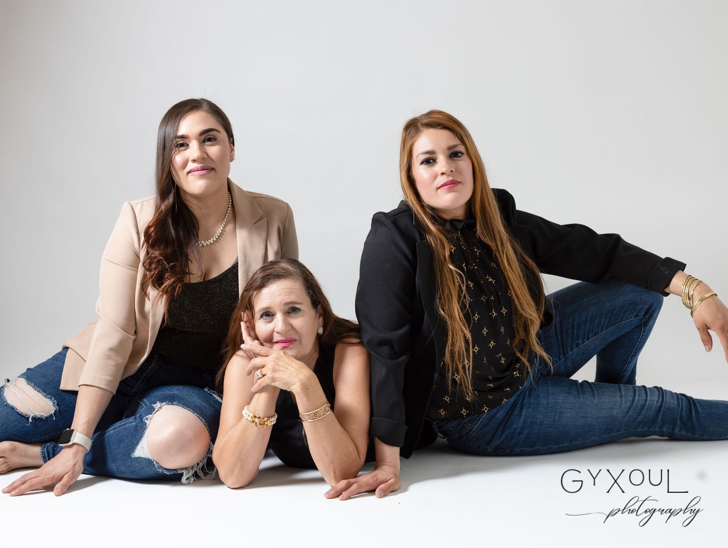 Three women pose casually together on the floor in a minimalist studio portrait.
