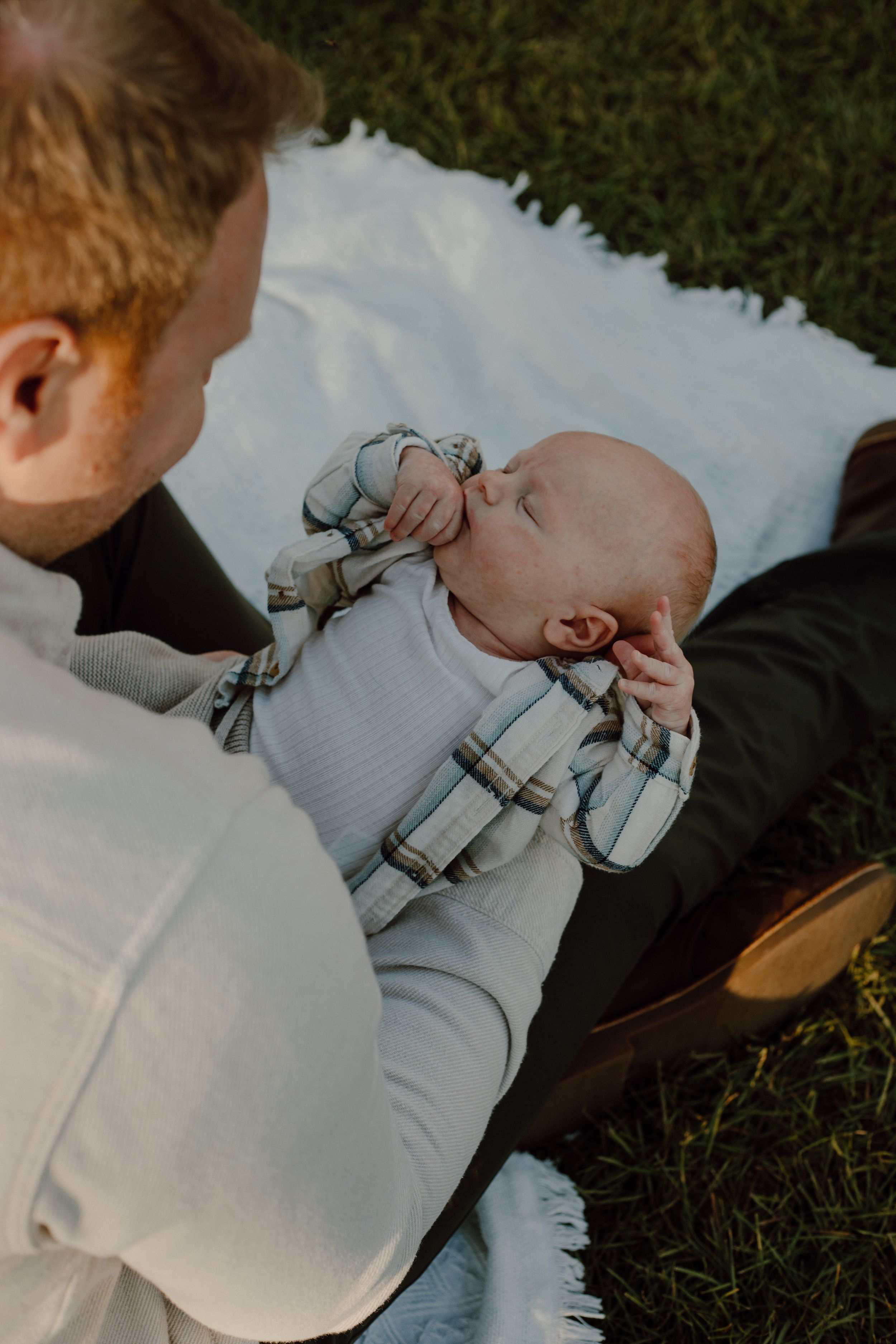 Baby in plaid outfit lying on white blanket outdoors during golden hour.