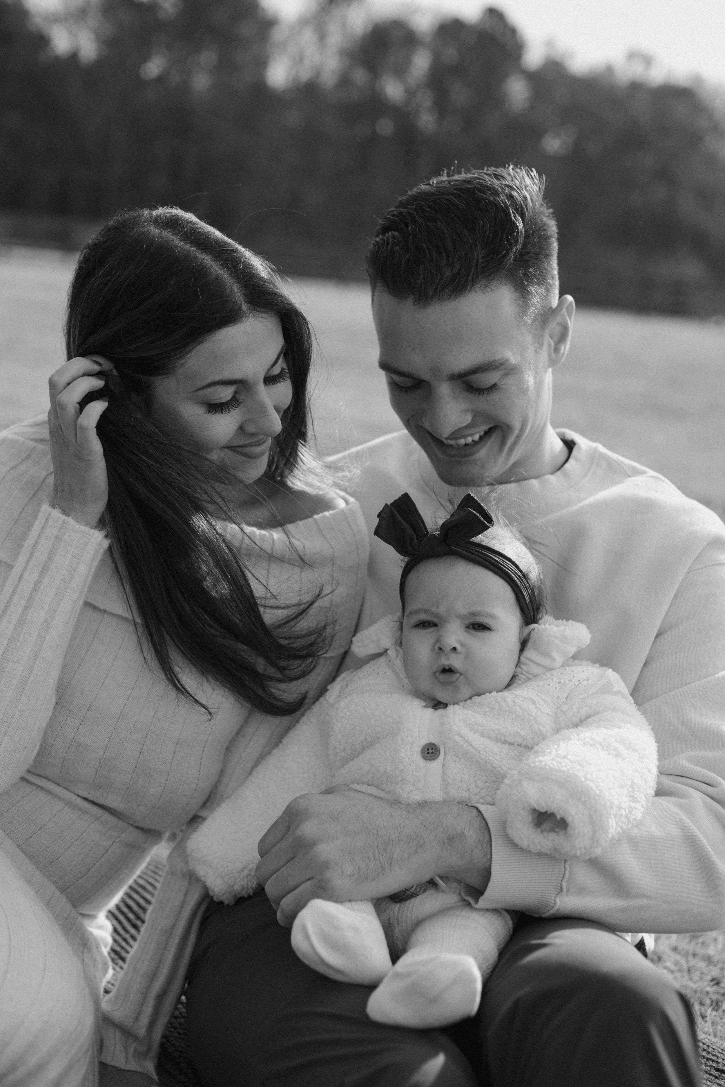 A heartwarming black and white family portrait showing parents lovingly embracing their baby wearing a bow outdoors.