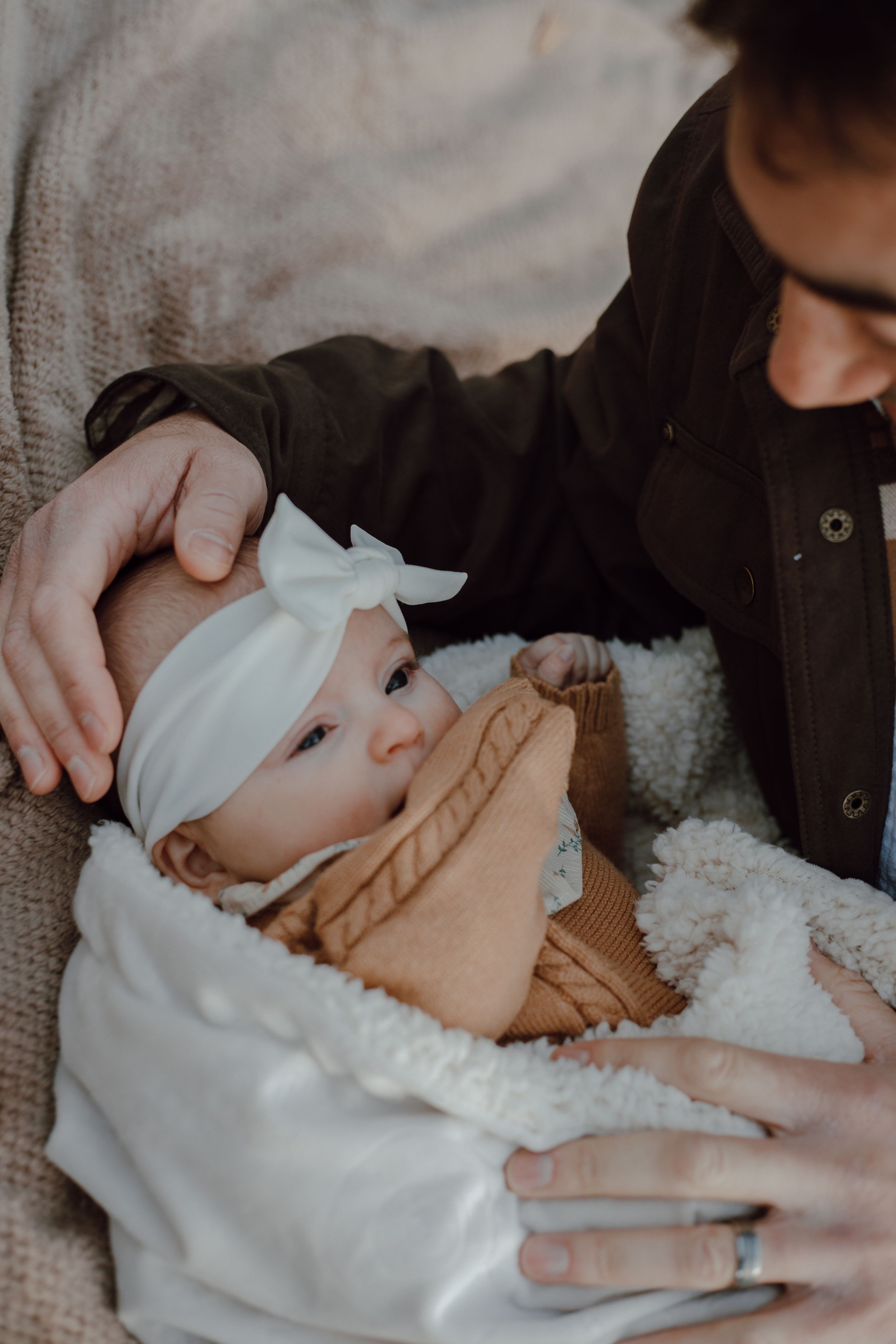 A baby wearing a white headband and tan outfit is being cradled in a cozy white blanket.