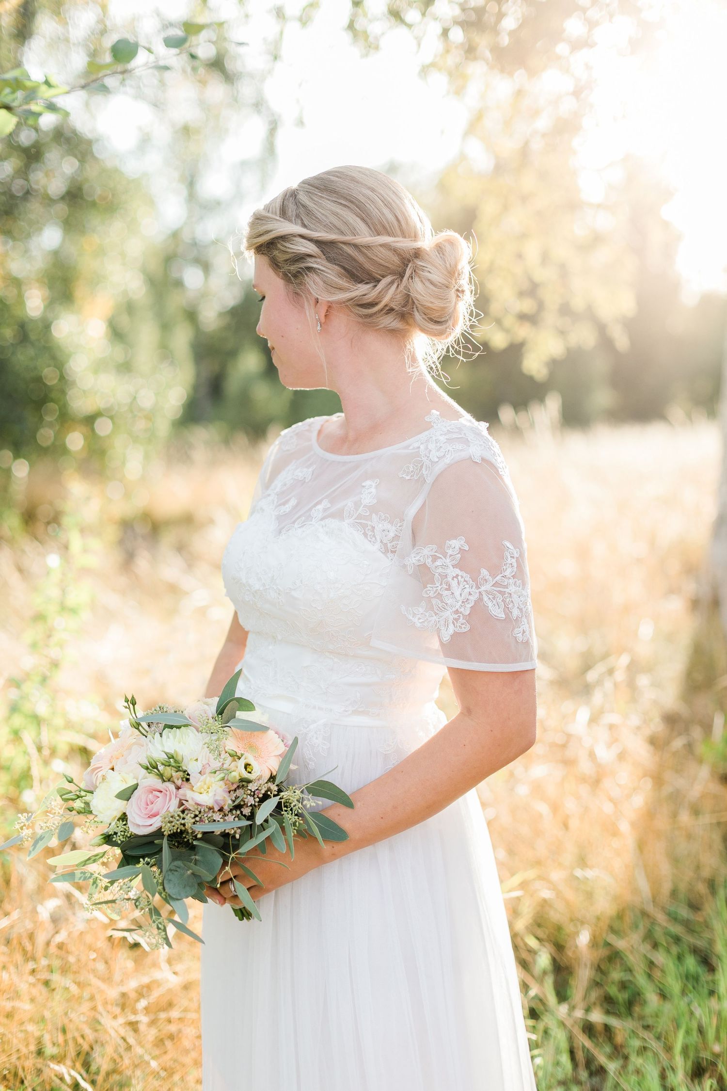 Bride in delicate lace dress holds pastel bouquet in golden sunlight.