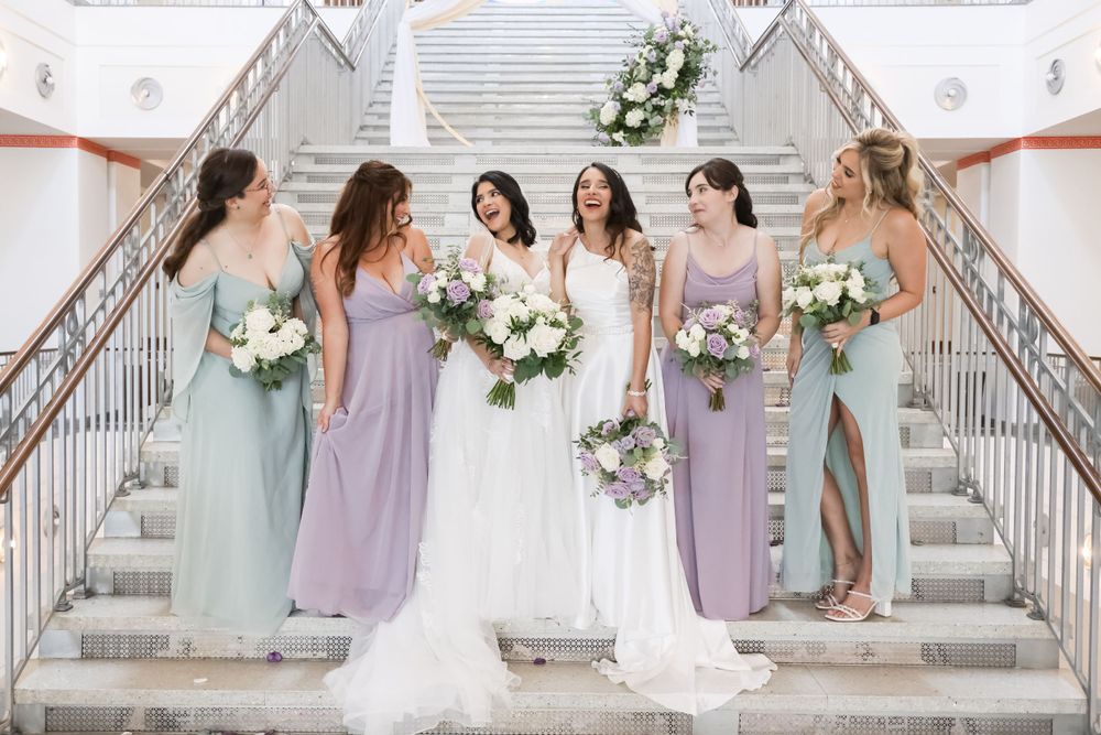 A bridal party in pastel purple and mint bridesmaid dresses poses on white marble stairs.
