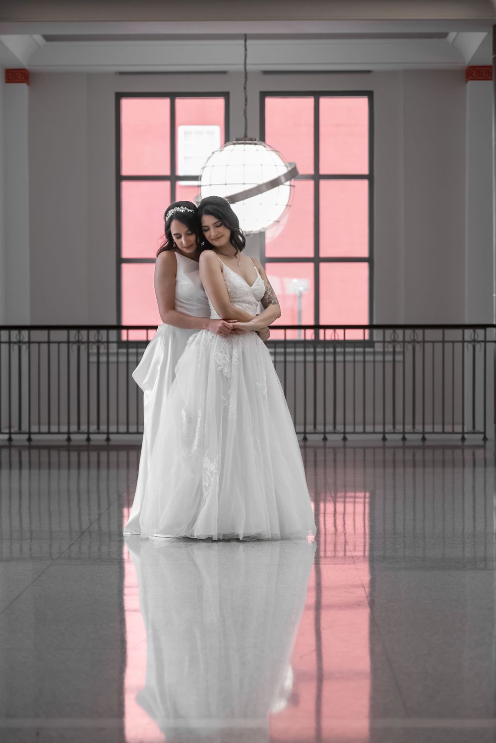Two brides in white gowns embrace in front of pink-lit windows with modern industrial decor.