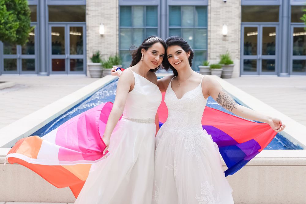 Two brides in matching white dresses pose with a colorful pride flag outdoors.