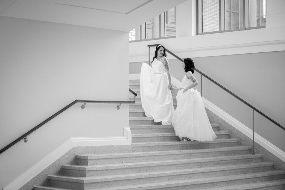 A black and white photo captures two people in wedding dresses descending a modern staircase.