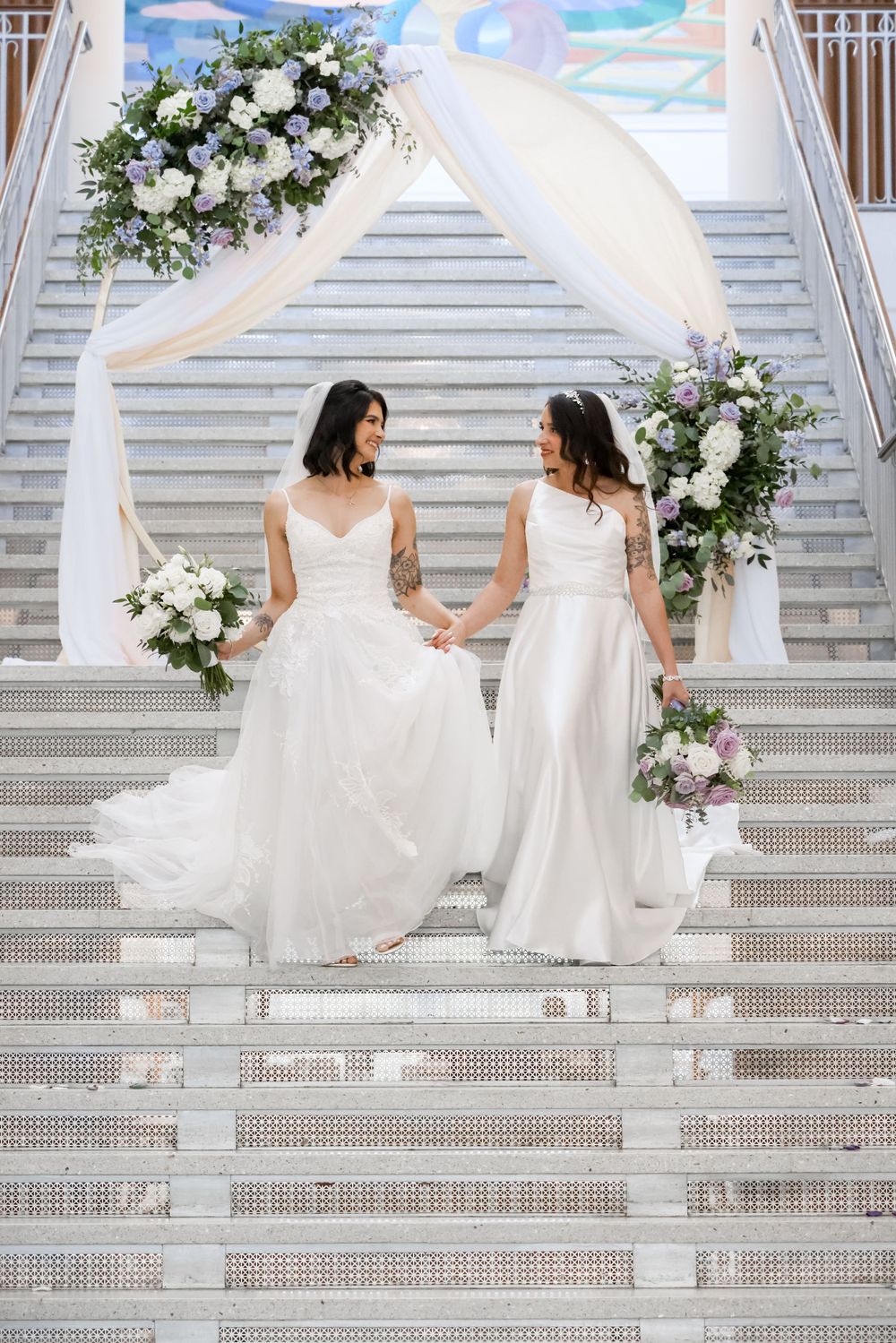 Two brides in white wedding gowns sitting on elegant white staircase decorated with flowers.