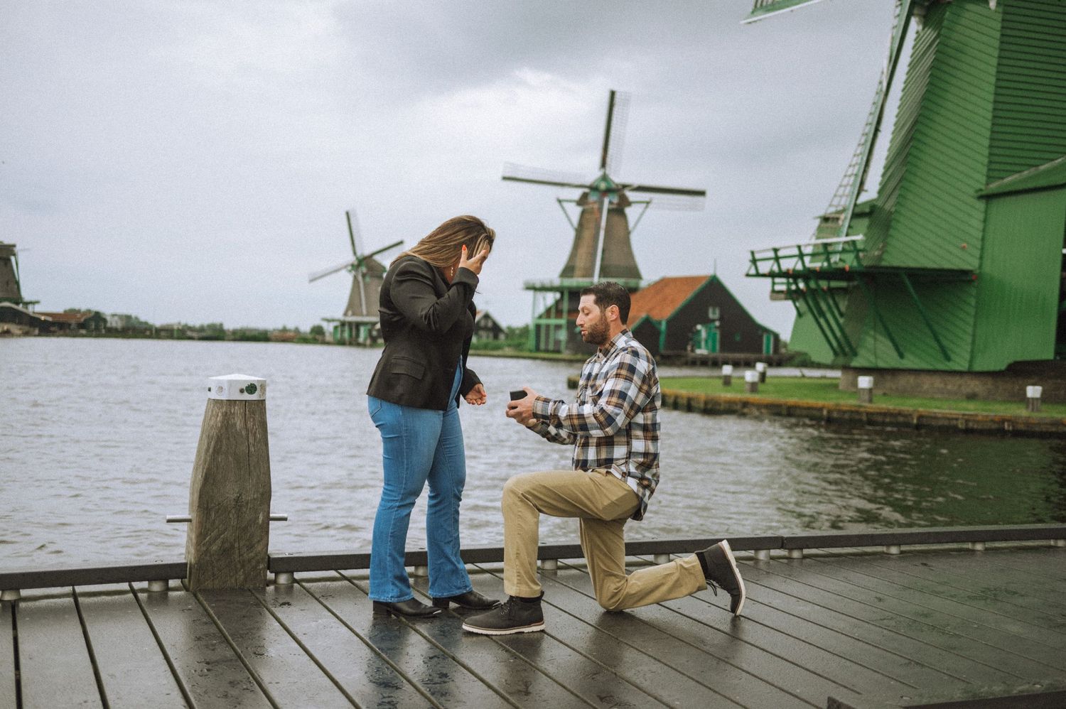 Secret proposal in Zaanse Schans – A man kneels before his surprised partner, with traditional Dutch windmills in the background.