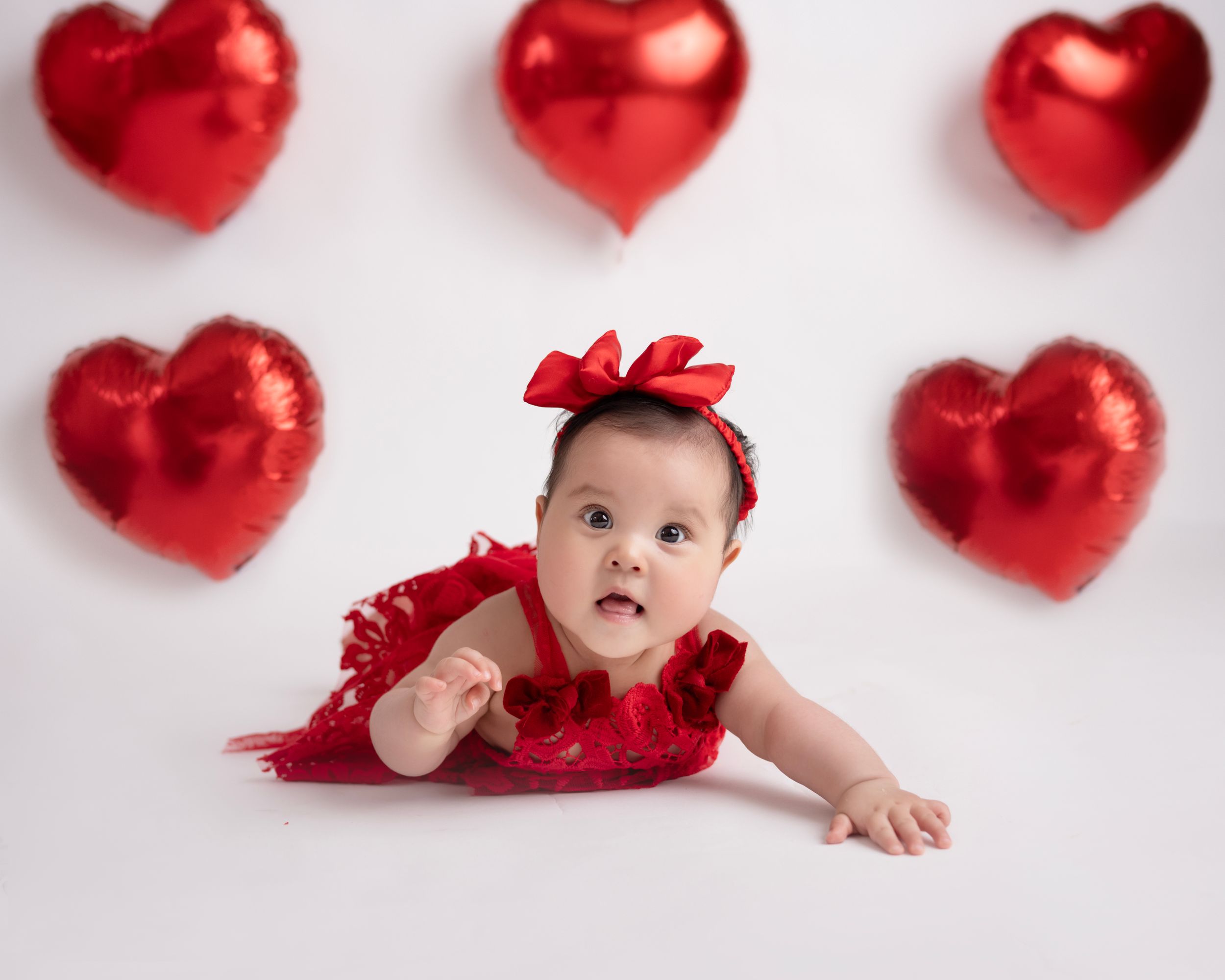 Baby in red outfit lying among shiny red heart decorations on white backdrop.