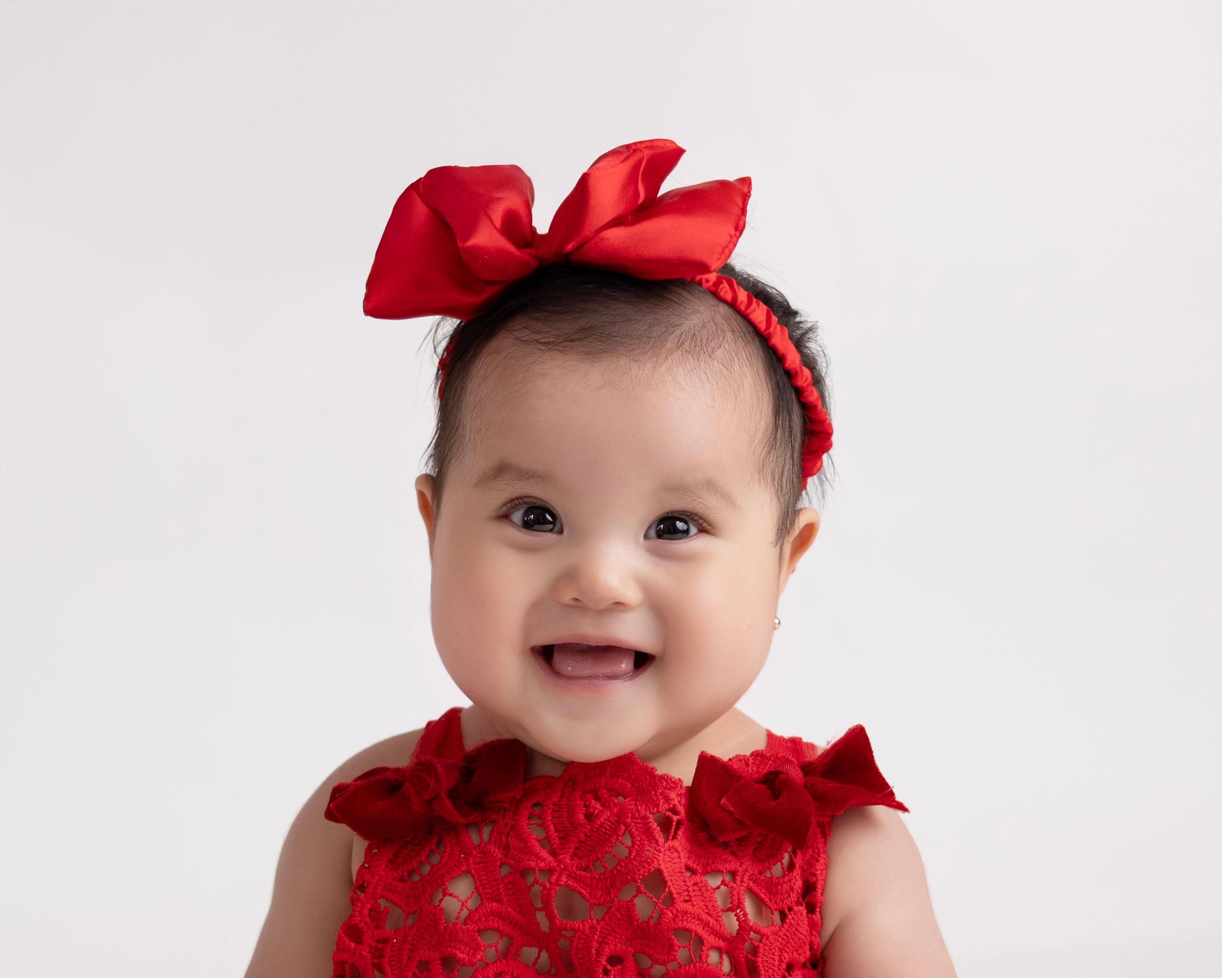 Closeup portrait of baby in red bow and dress against white background with heart decorations.