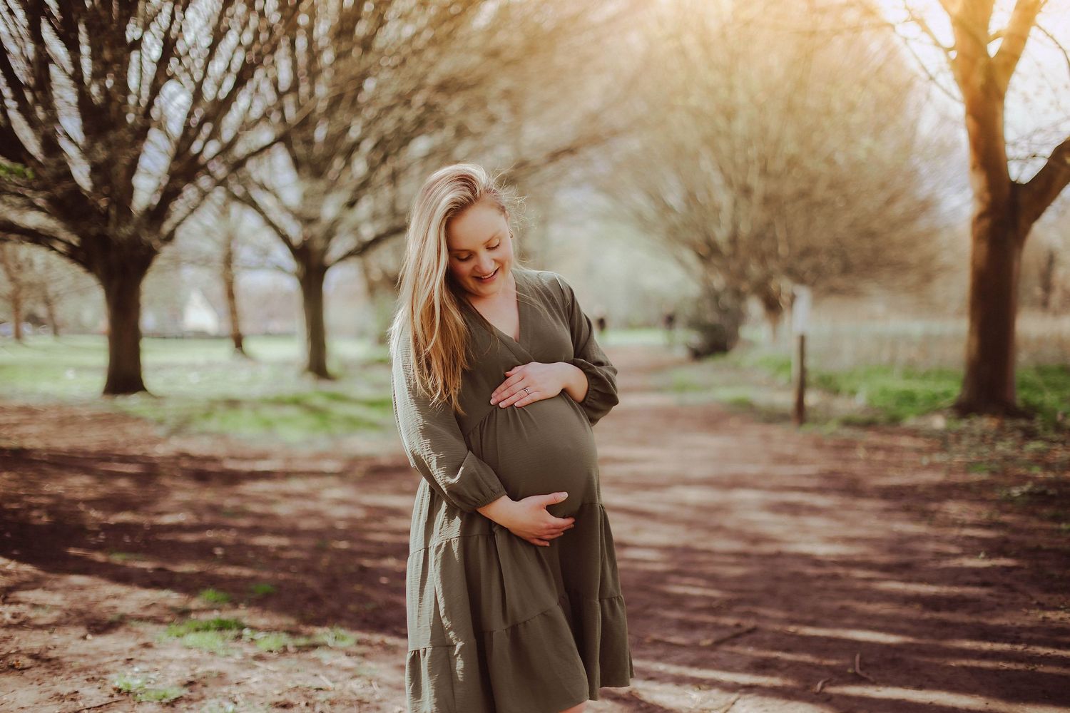 Expectant mother in a flowing gray dress stands on an autumn path beneath blooming trees at sunset.