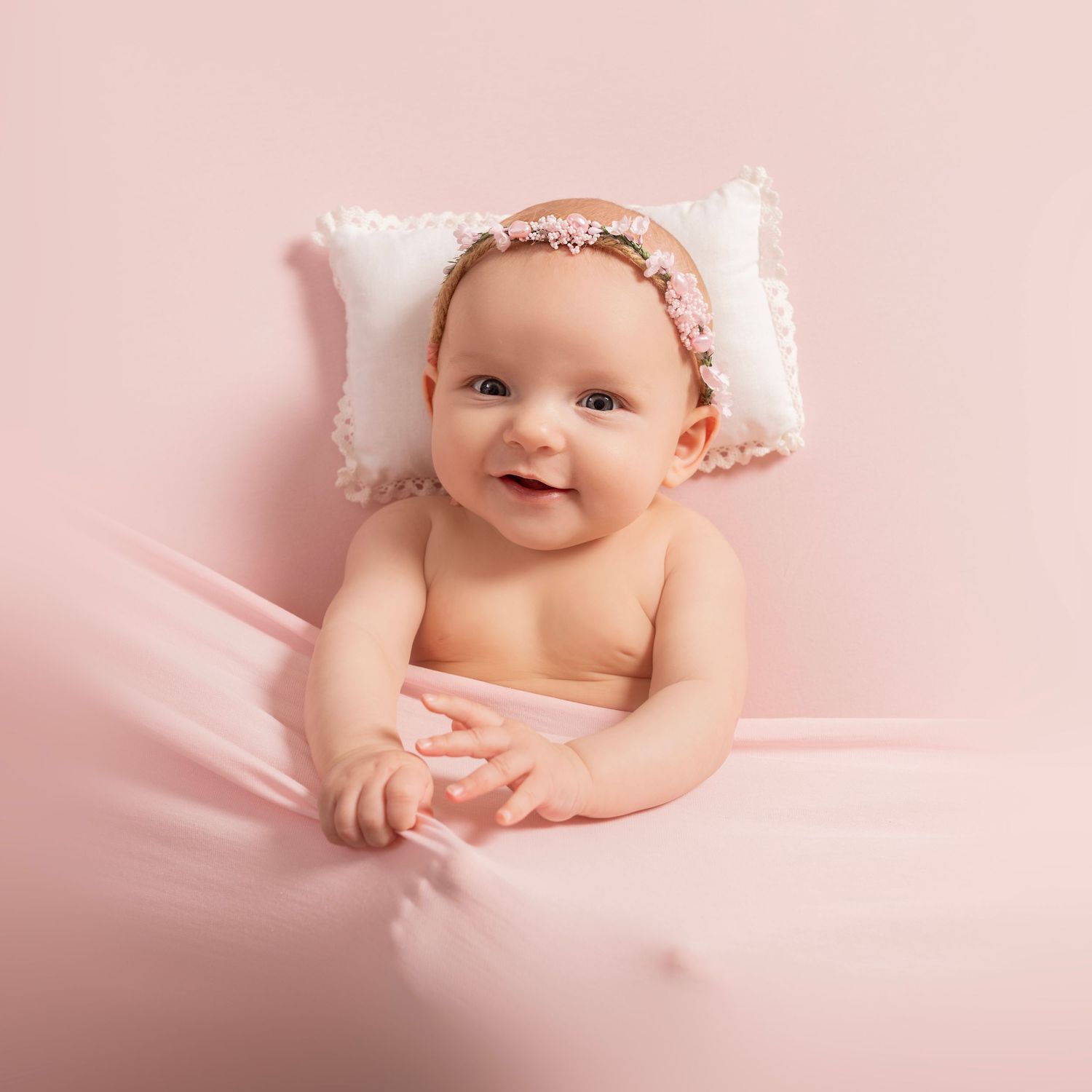 A baby wearing a pink headband lies on soft pink fabric in a gentle portrait pose.