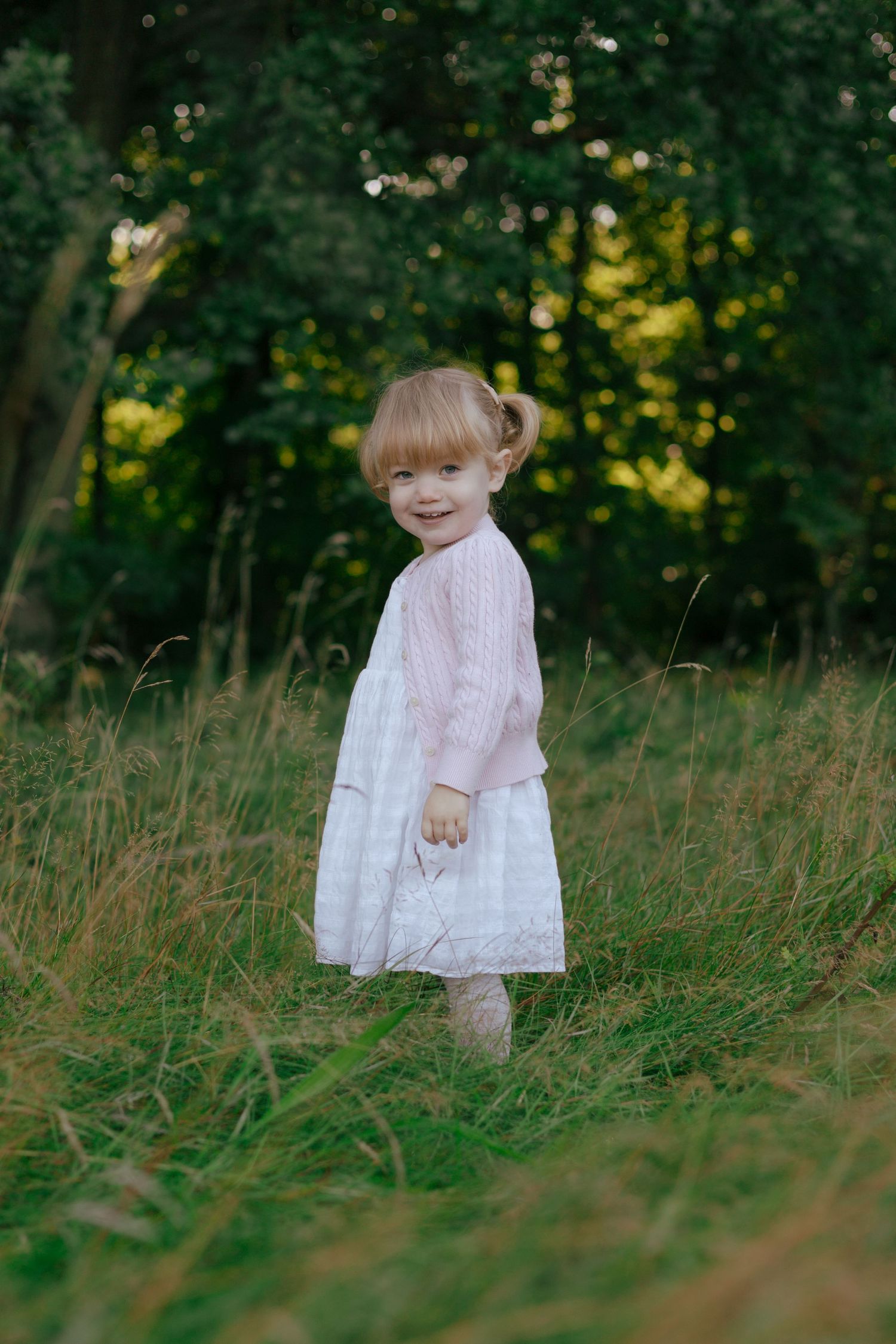 Child in pink and white dress standing in tall grass with sunlit forest behind.