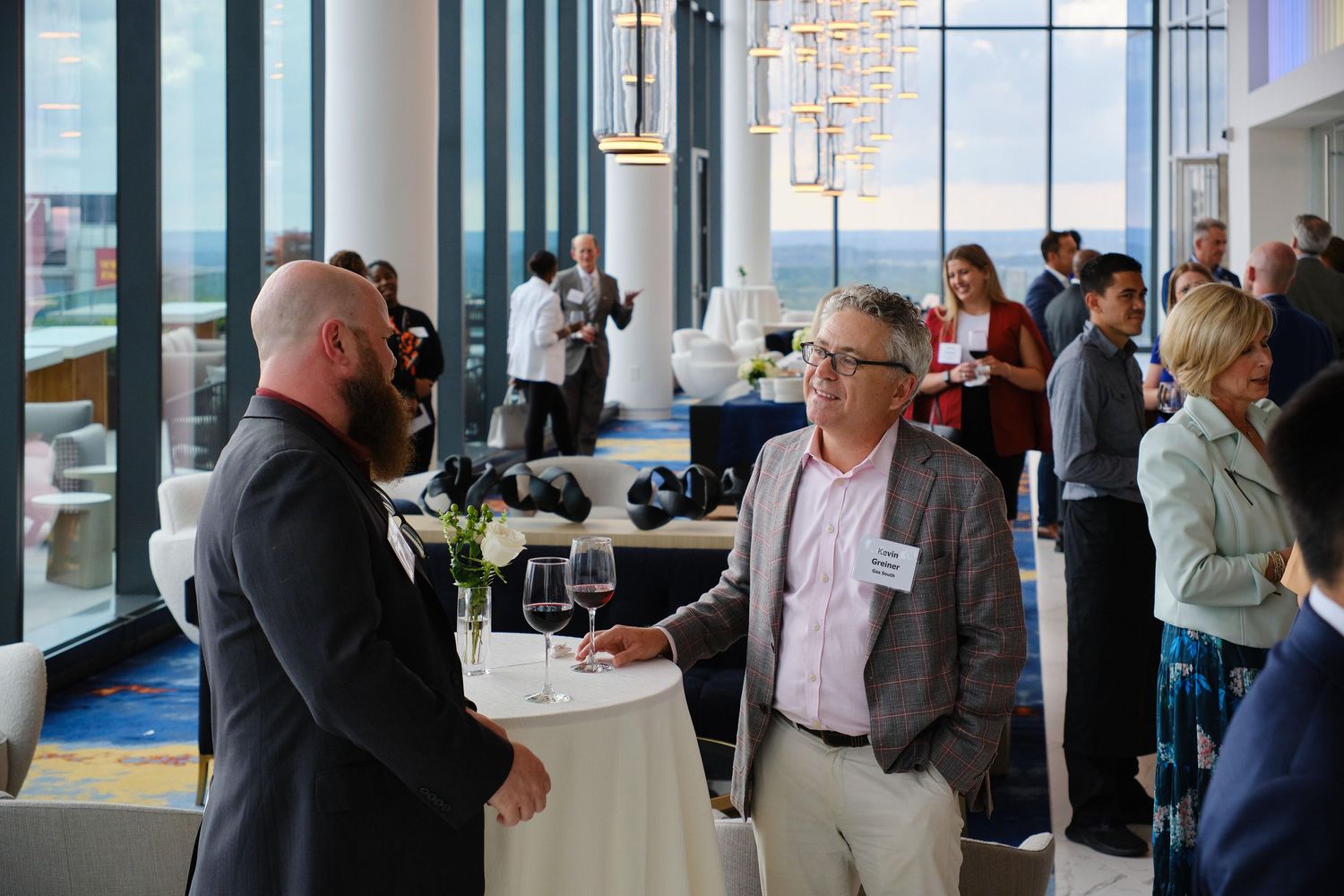 Business professionals networking at an upscale event venue with floor-to-ceiling windows and cocktail tables.