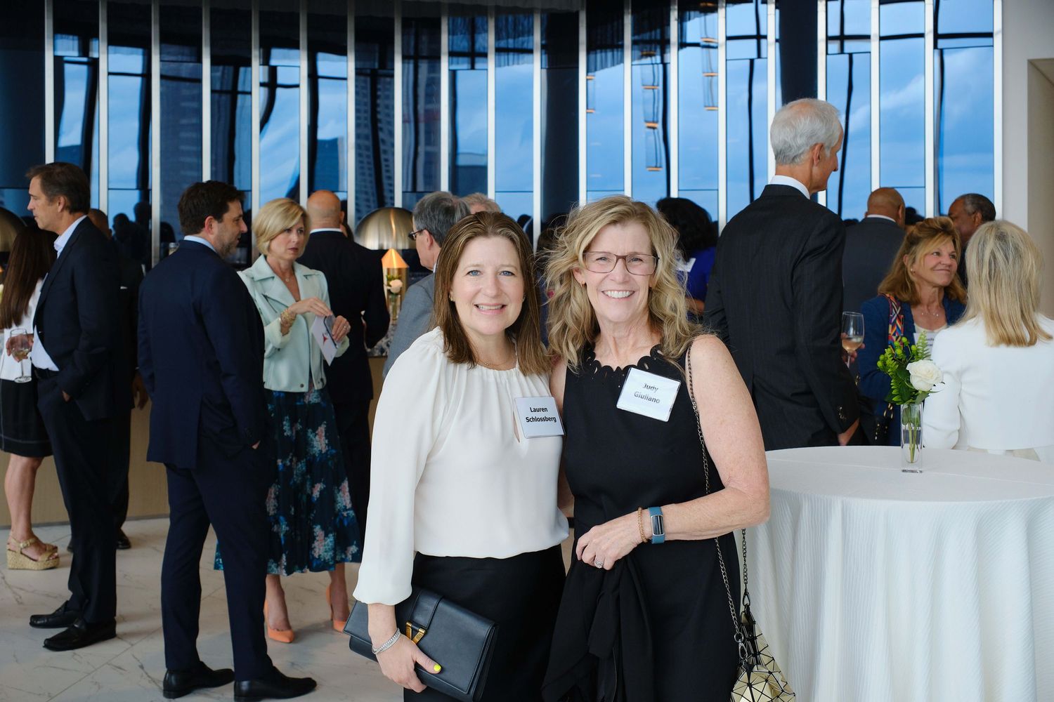 Professional networking event in a modern venue with attendees mingling near tall windows.