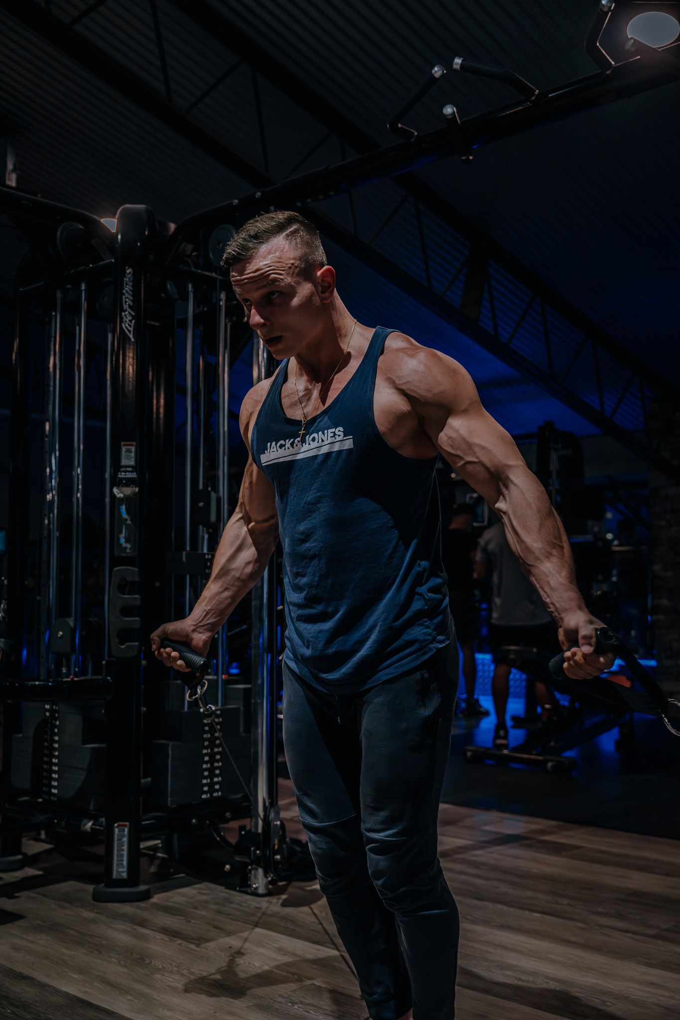 Muscular athlete performing cable exercises in a dark gym wearing a navy tank top and sunglasses.