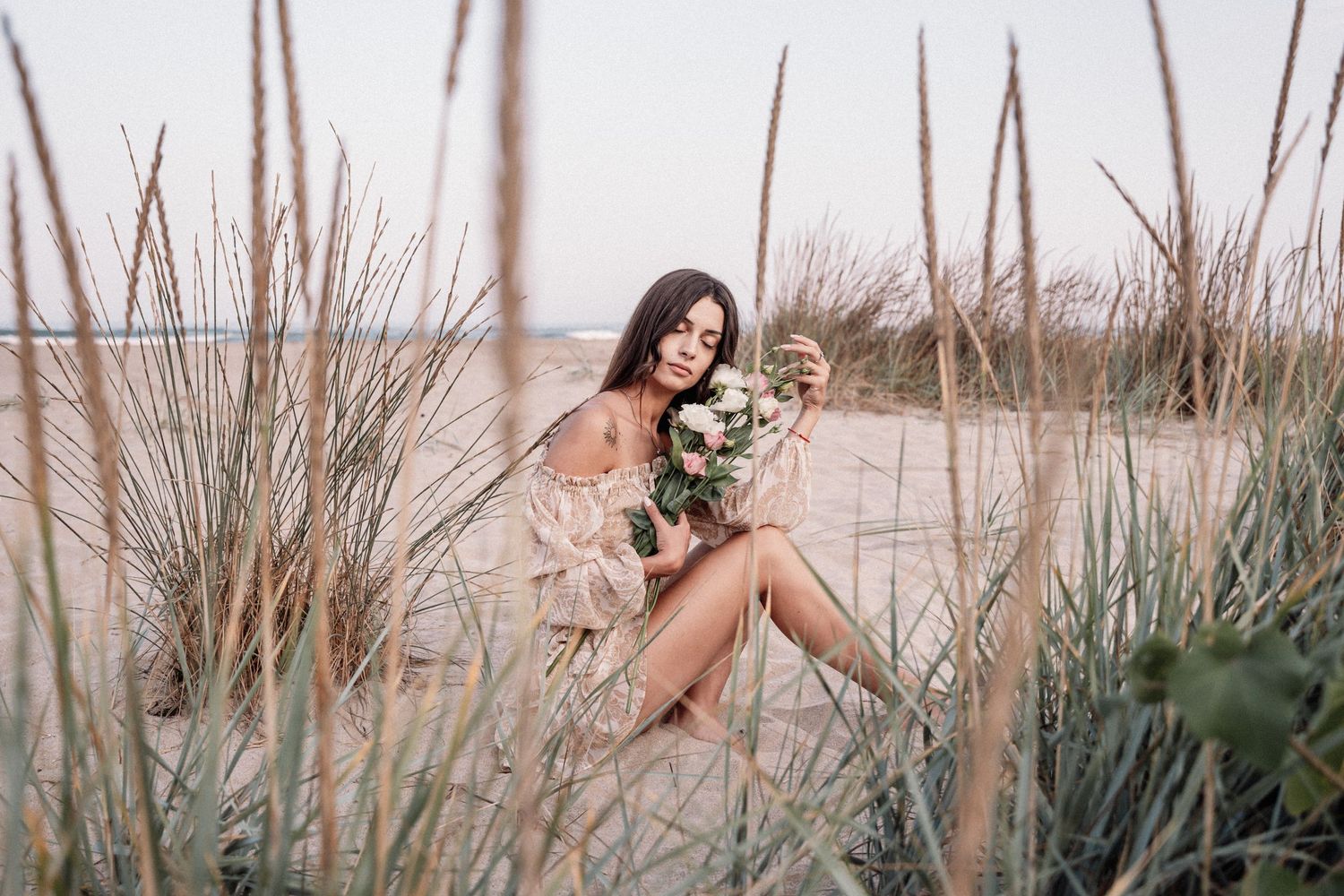 Woman in pink dress sits among tall beach grass holding floral bouquet.