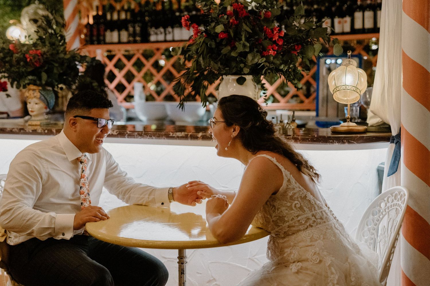 A couple in elegant wedding attire shares an intimate moment at a table in a warmly lit venue.