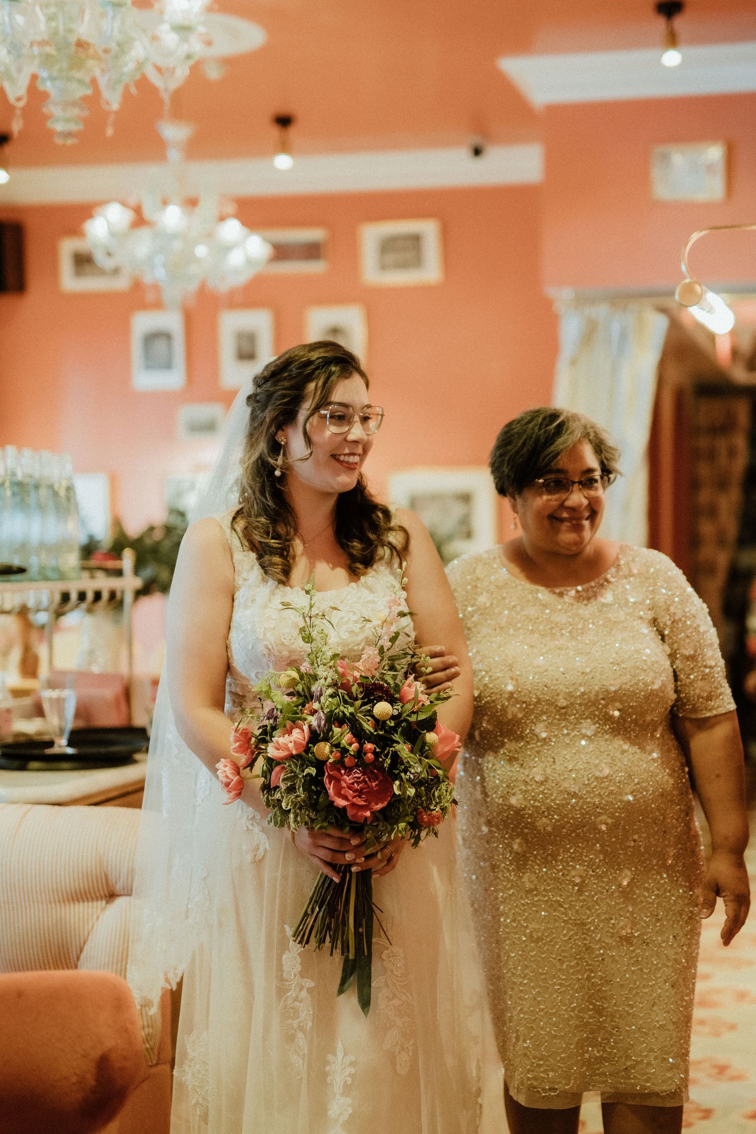 A bride in a white lace dress stands with an older woman in gold sequin dress holding a colorful bouquet in a pink room.