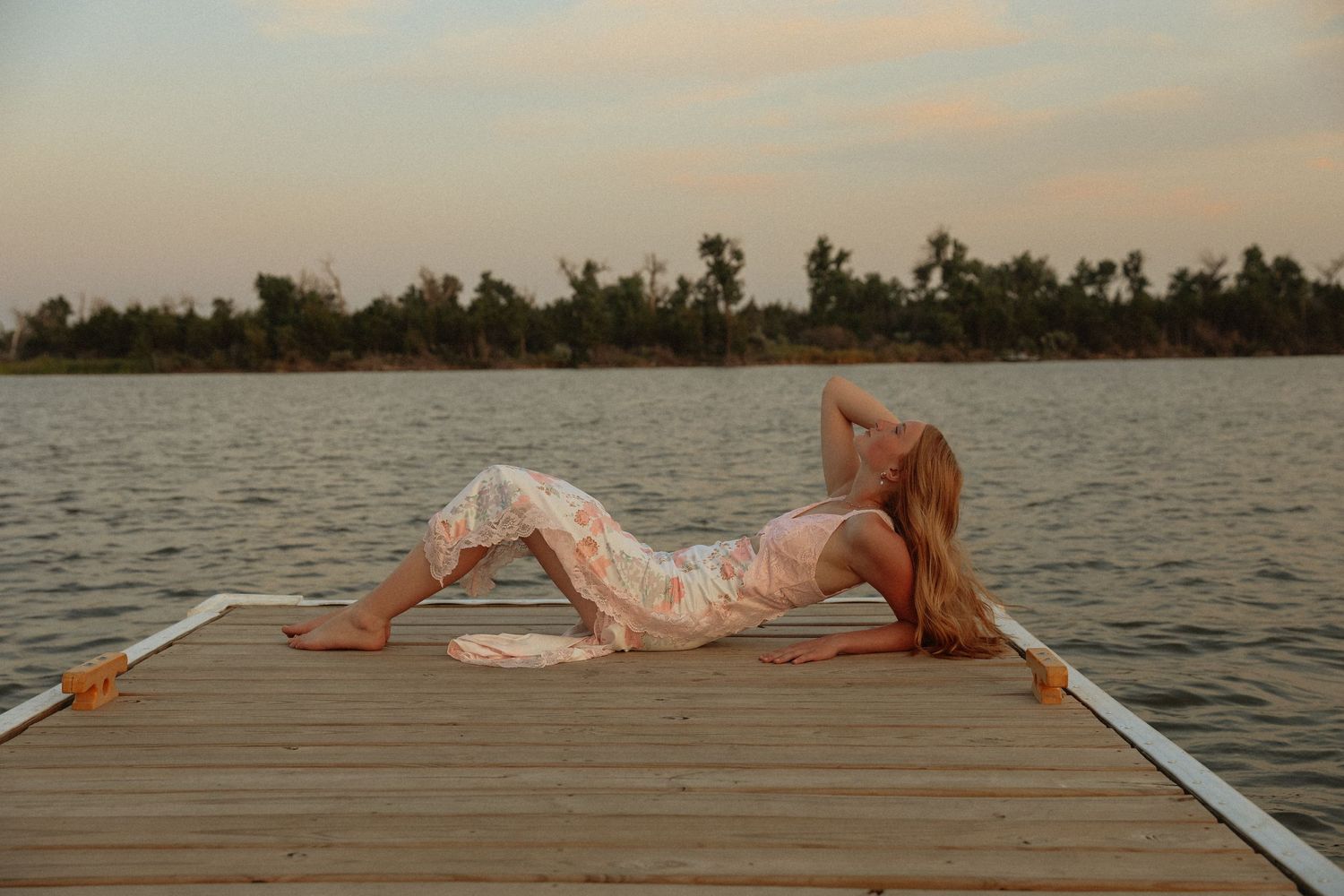 A person lies stretched out on a wooden dock at sunset over calm waters.