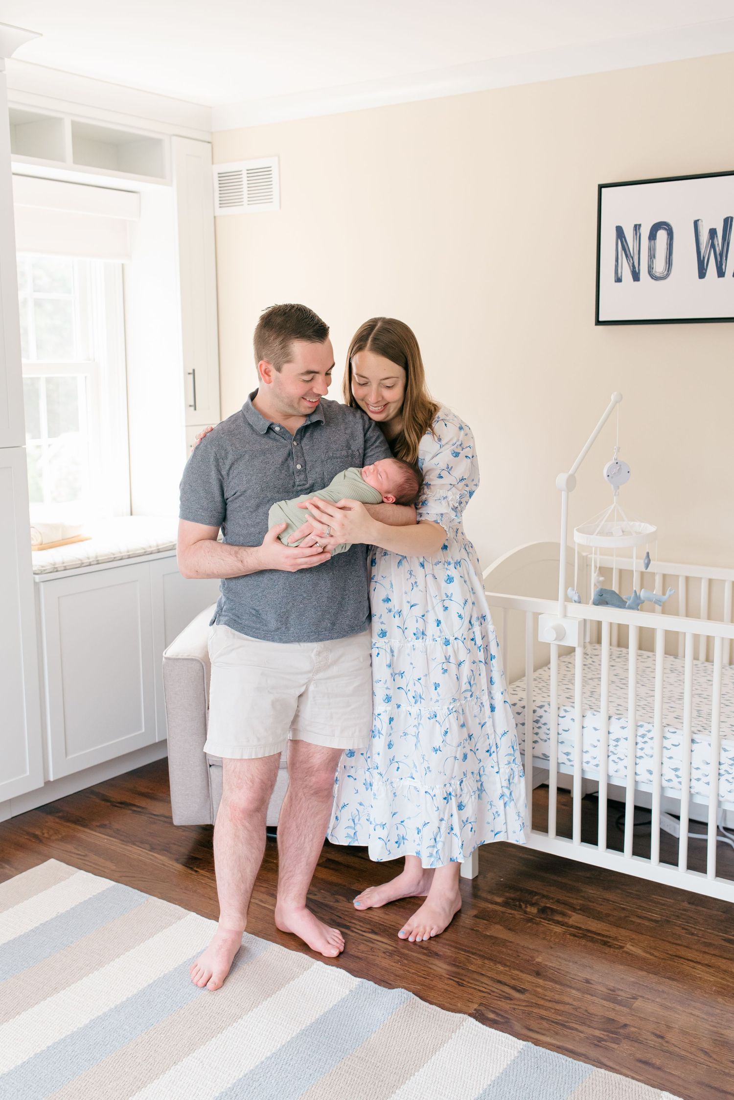 A man and woman hold their newborn baby in a nursery with beige walls and a white crib.