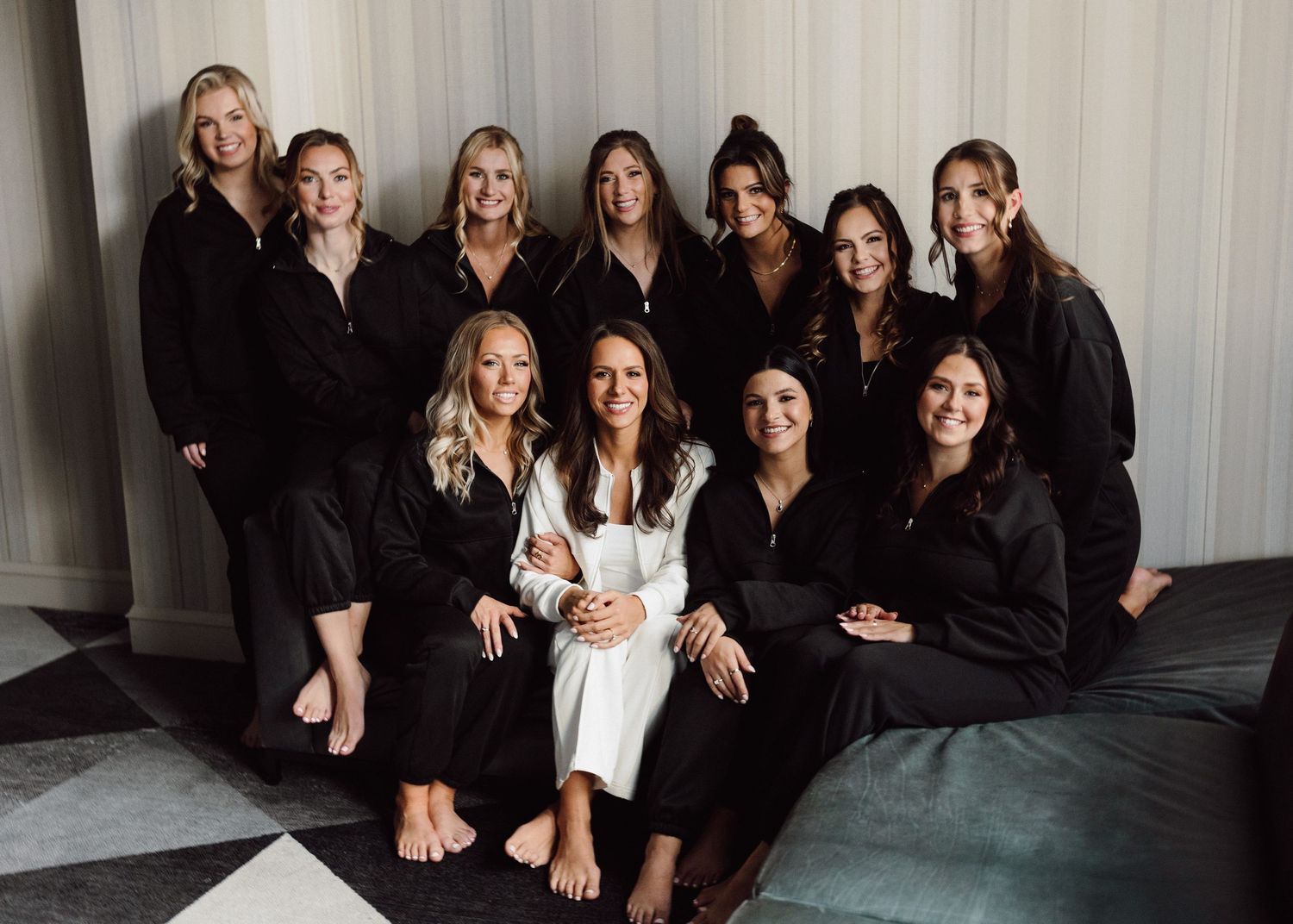 Group photo of bridesmaids in black robes gathered around bride in white on a couch.