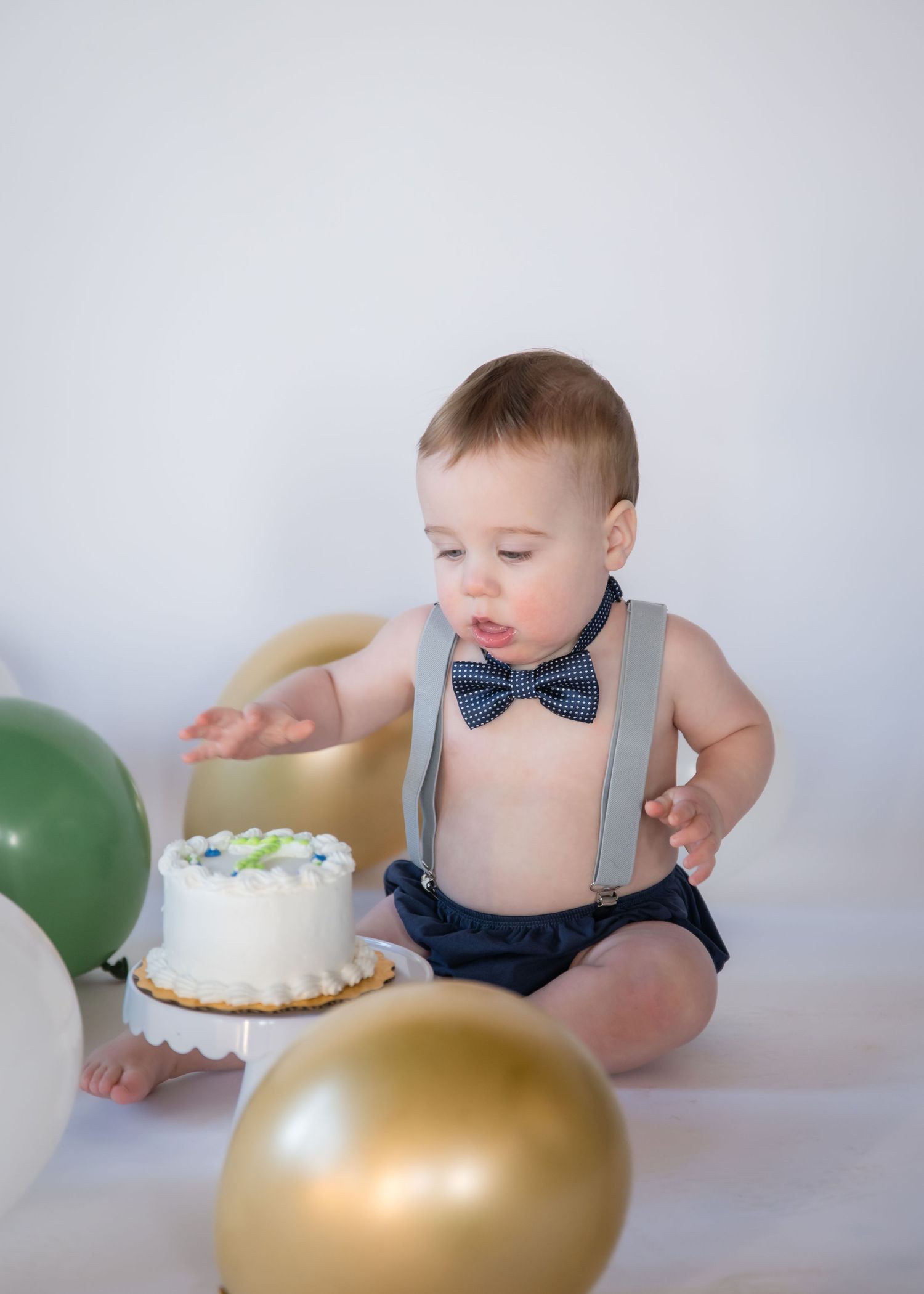 Sweet cake smash portrait features baby in suspenders and bow tie with white cake and gold balloon decorations.