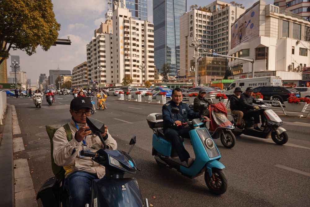 Commuters on scooters and motorbikes wait at intersection in urban city street with tall buildings in background.
