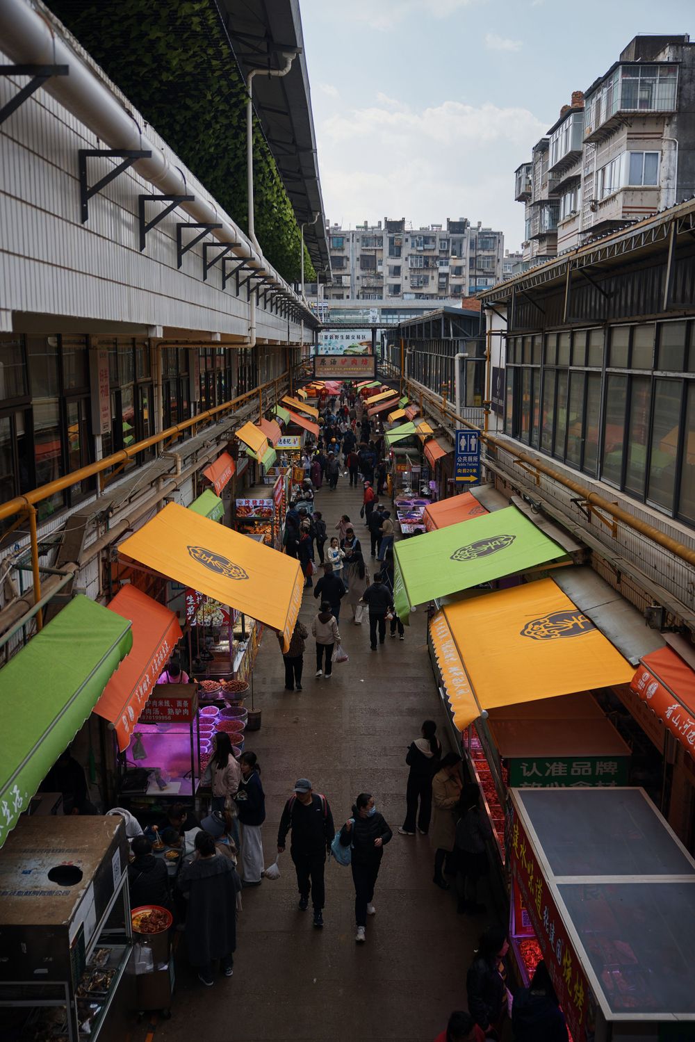 Bustling marketplace alley with colorful green and orange vendor stalls beneath covered walkway in urban residential area.