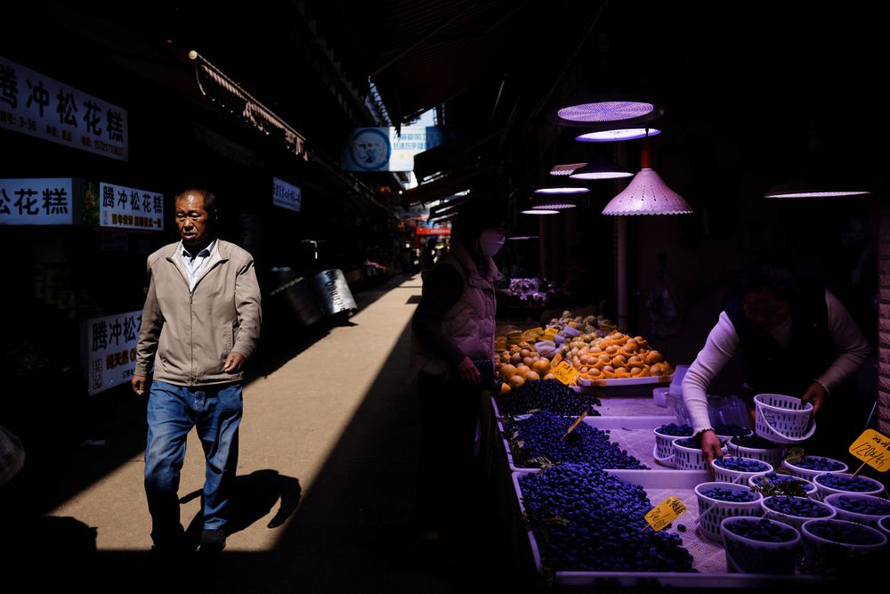 A person in a beige jacket walks through dramatic lighting in a market with produce displayed on the right.