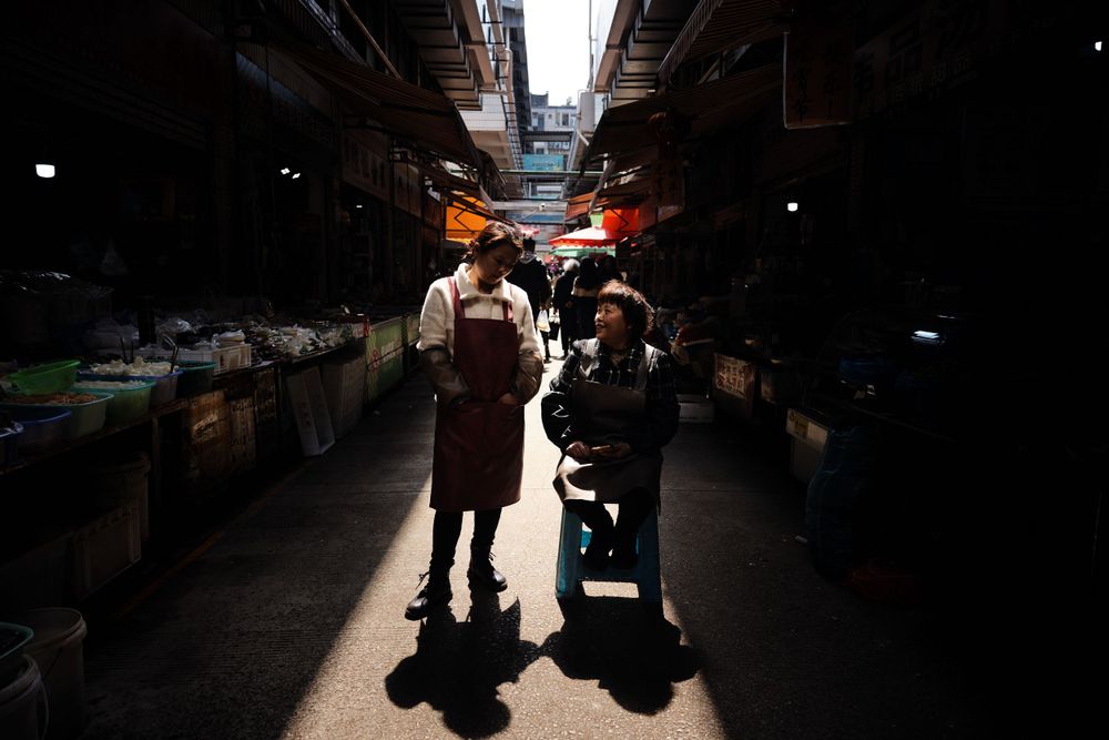A silhouetted pair of people walking through a dark indoor market alley with light streaming from above.