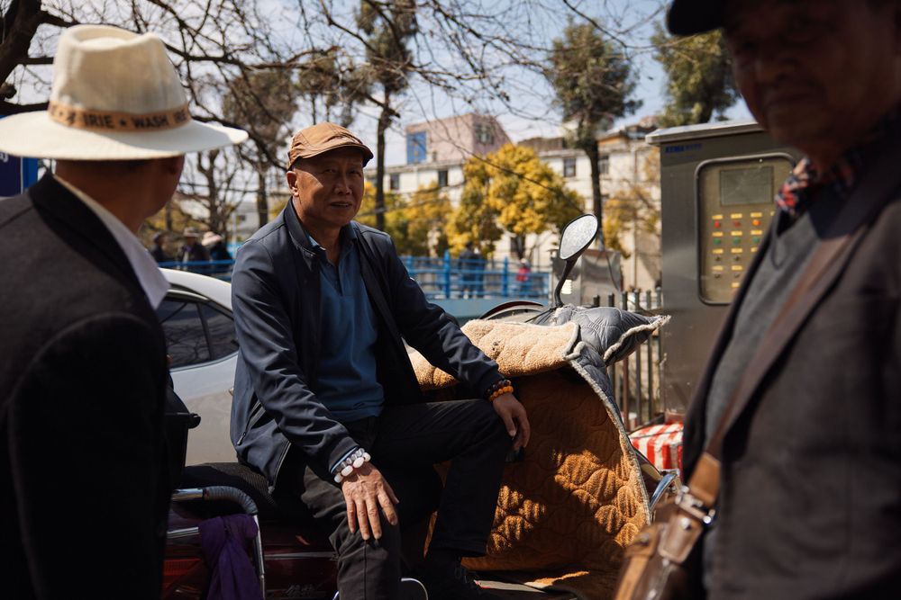 People gather around a market stall or vendor table on a sunny day with trees and buildings in the background.