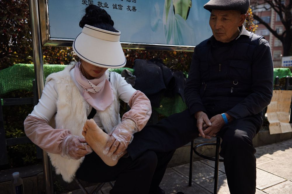 Two people sitting outdoors, one in a white sun visor and pink jacket, another wearing a dark cap and coat.